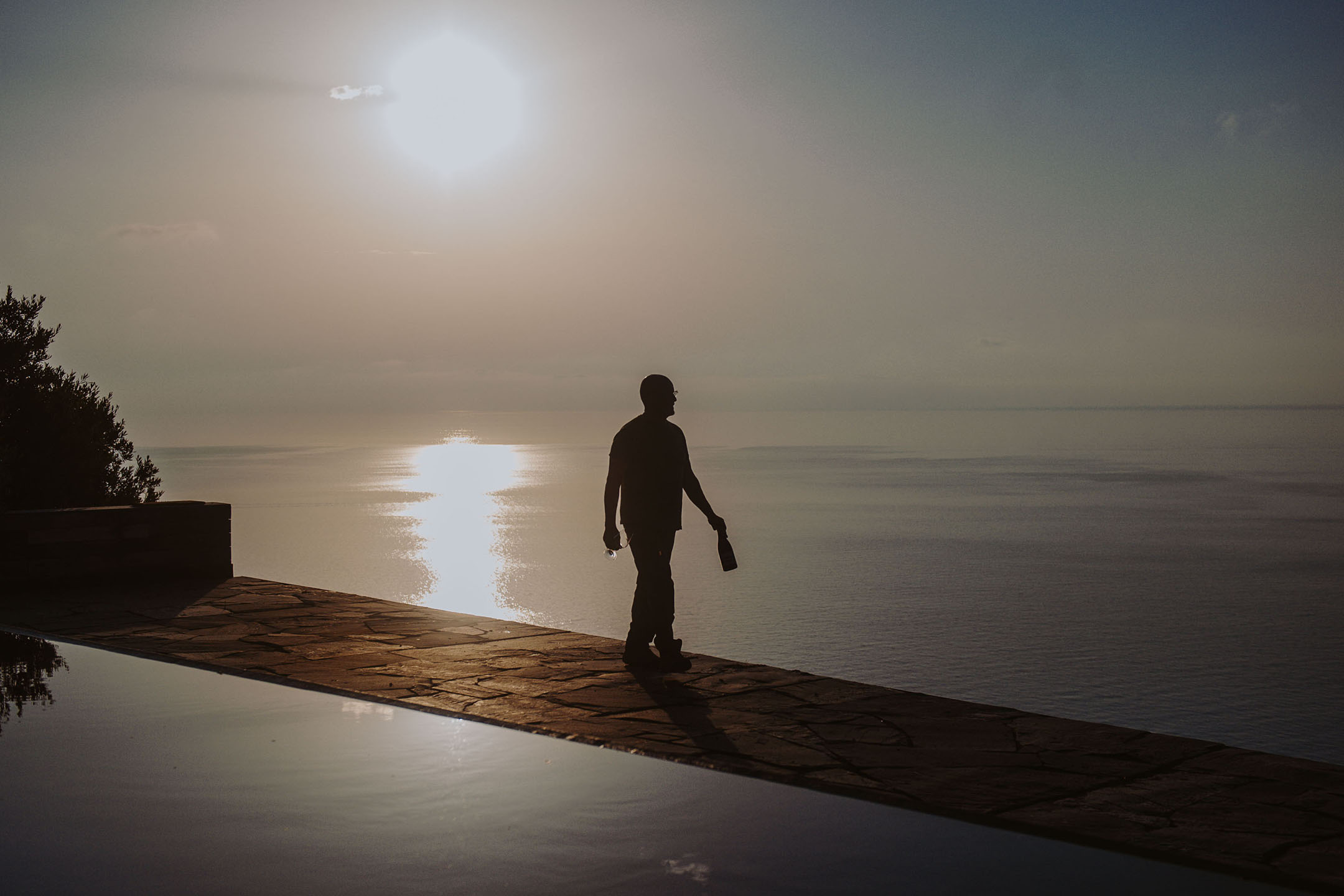 Silhuett av en person med flaska och glas vid en infinitypool vid solnedgången