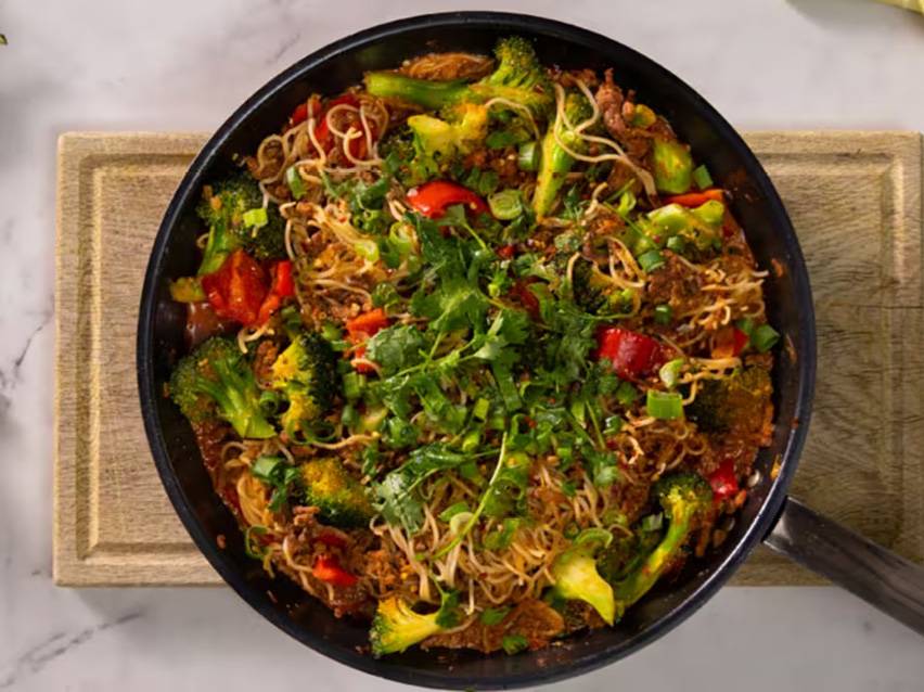 En stekpanna med nudlar, broccoli, rött paprika, kött och färska örter.