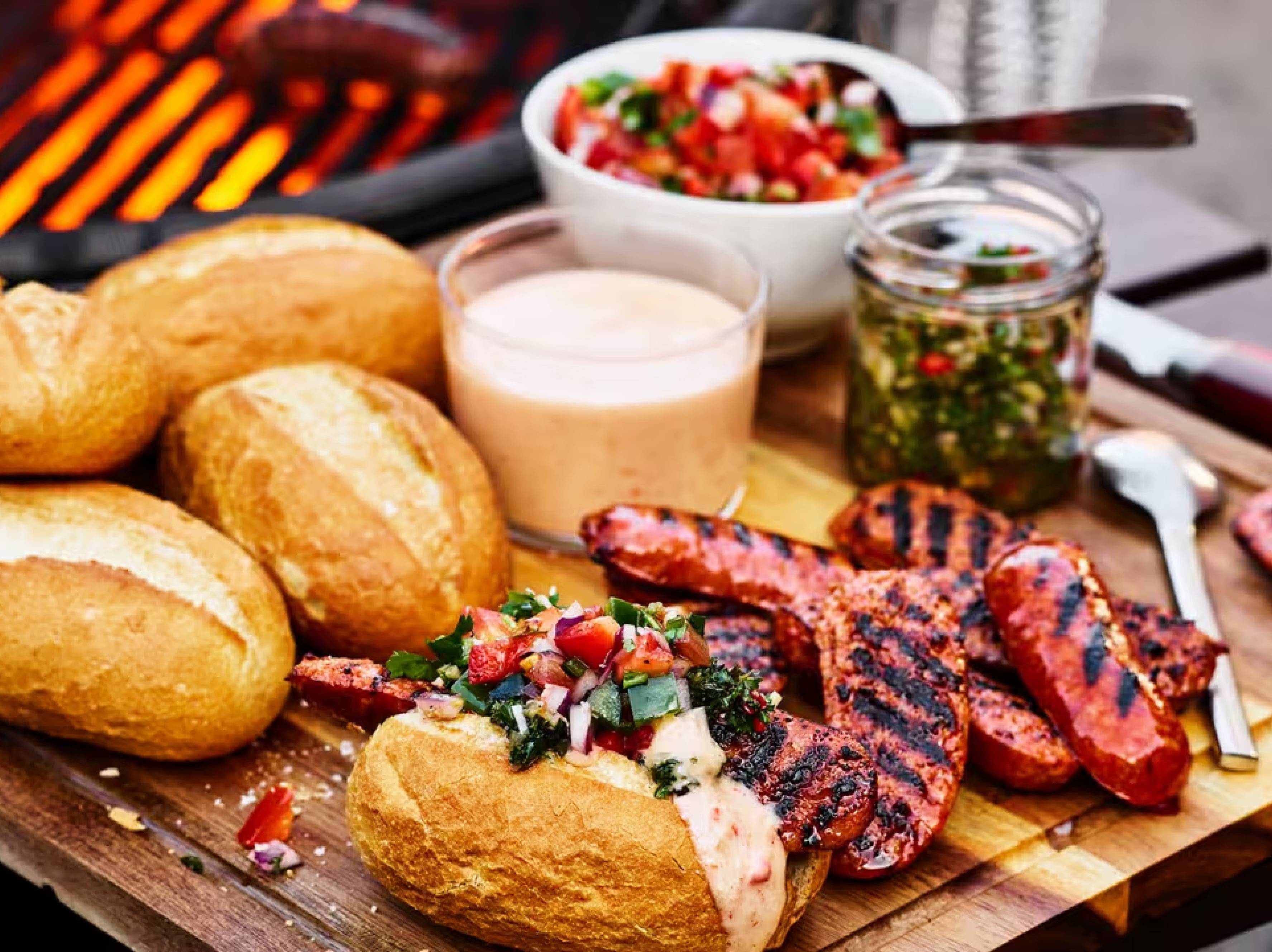 Grillade korvar och bröd med salsa och sås på en träskärbräda, med en grill i bakgrunden.