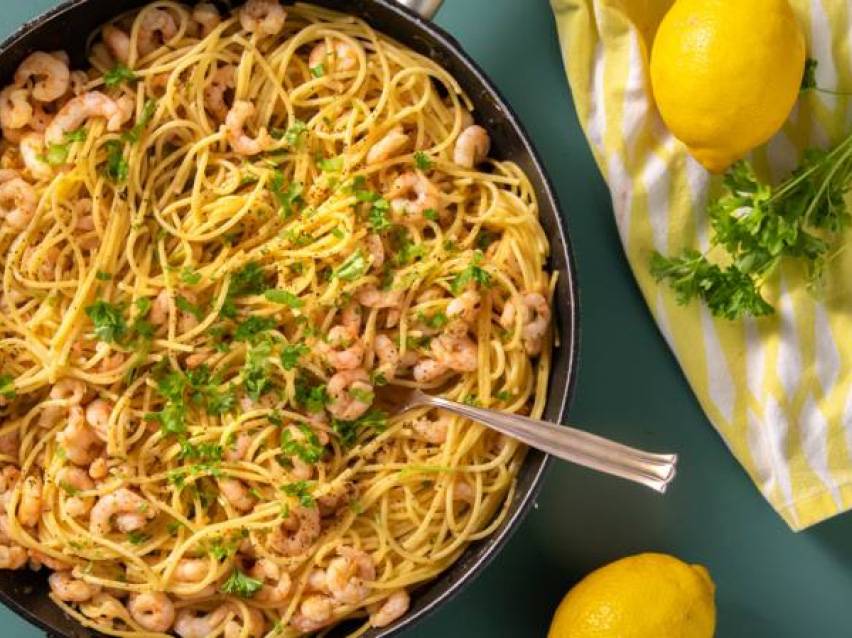 Spaghetti med räkor och persilja i en panna, med citroner och en kökshandduk bredvid.