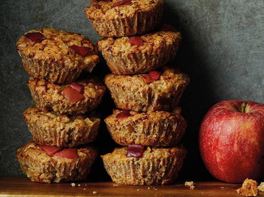 En hög med äppelmuffins med äppelbitar på toppen, bredvid ett rött äpple.