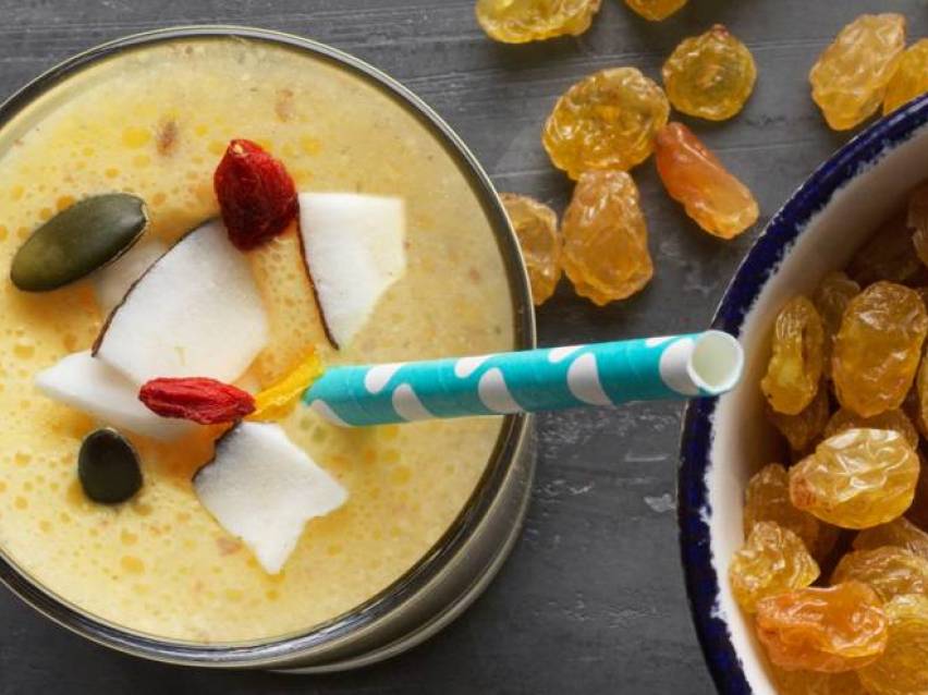 Gul smoothie med kokos, gojibär och pumpafrön, bredvid en skål med russin.