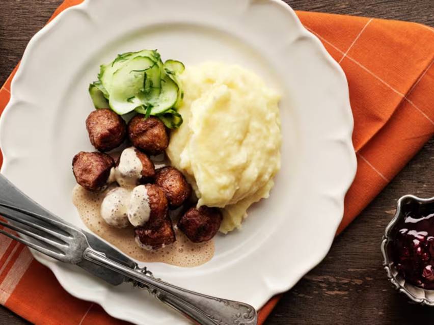 Köttbullar med gräddsås, potatismos, inlagd gurka och lingonsylt på en vit tallrik.