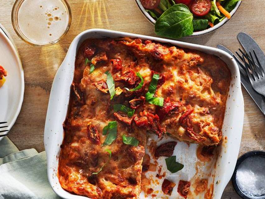 En ugnsform med lasagne toppad med soltorkade tomater och basilika, bredvid en sallad och ett glas öl.