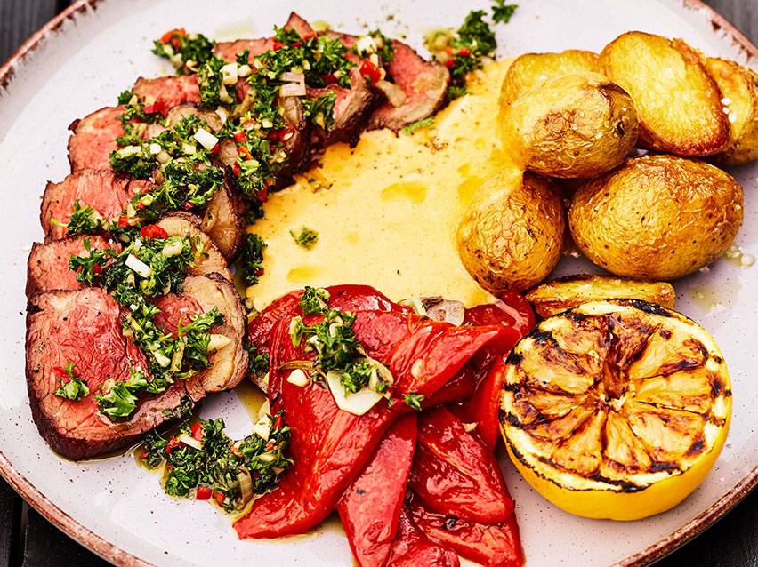 Skivad biff med chimichurri, potatis, grillad paprika och citron på en tallrik.