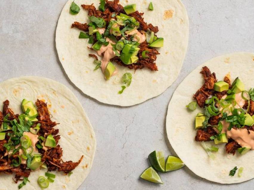 Tre tacos med strimlat kött, avokado, gräslök och sås, med limeklyftor.