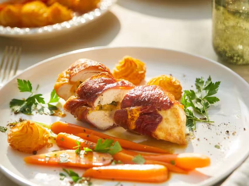 Kycklingfilé inlindad i bacon med morötter och potatispuré på en vit tallrik.