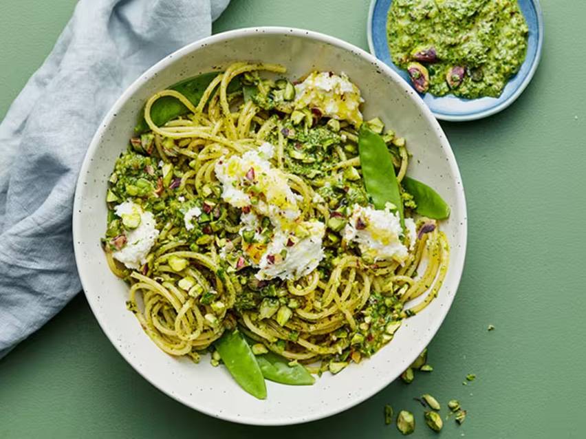 Spaghetti med grön pesto, pistagenötter, snöärtor och färskost i en vit skål.