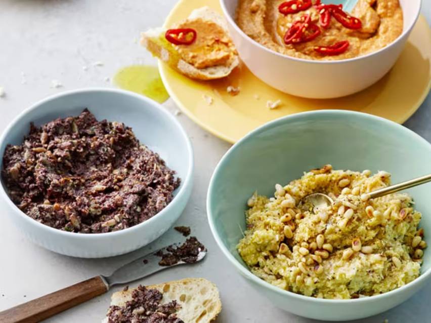 Tre skålar med olika dippar: tapenade, hummus och en grön dipp med pinjenötter, serveras med bröd.