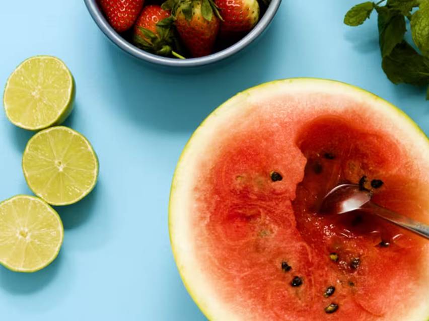 En halverad vattenmelon med en sked, skivade limefrukter och en skål jordgubbar på ett blått underlag.