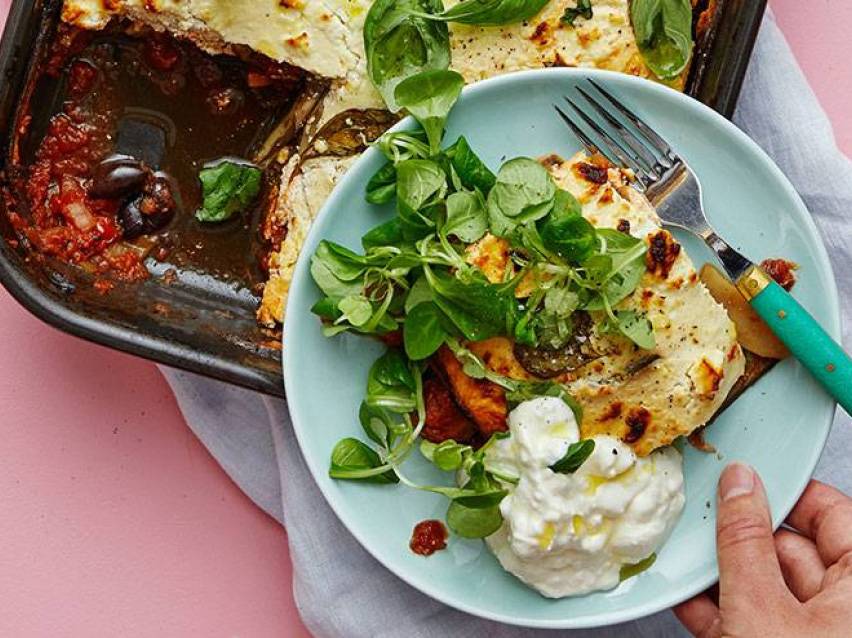 En portion moussaka med sallad och gräddfil på en tallrik, bredvid en ugnsform.