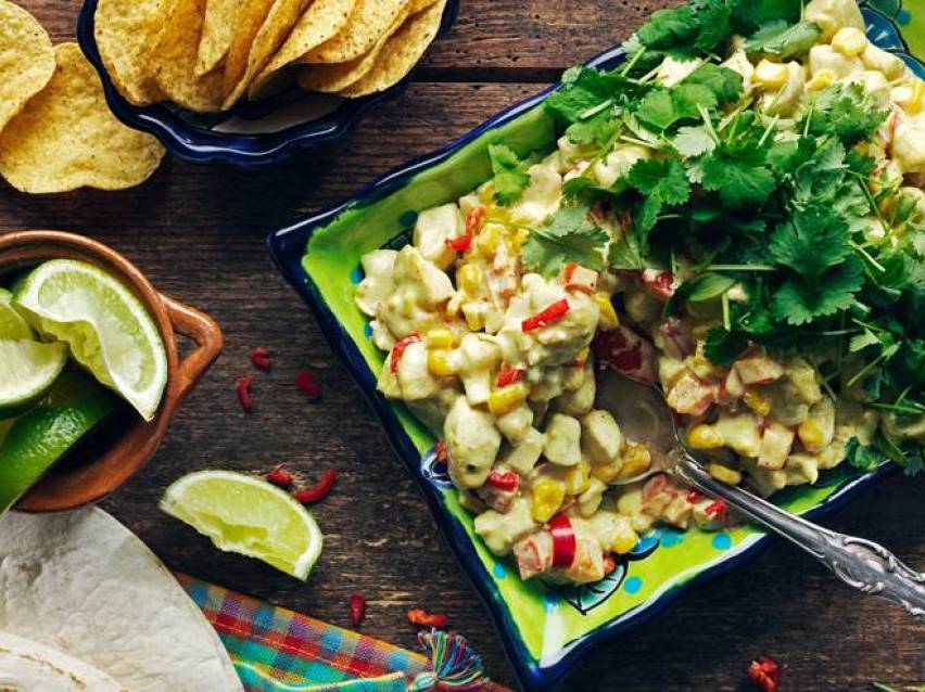 En skål med kycklingsallad med majs och paprika, toppad med koriander, bredvid tortillachips och limeklyftor.