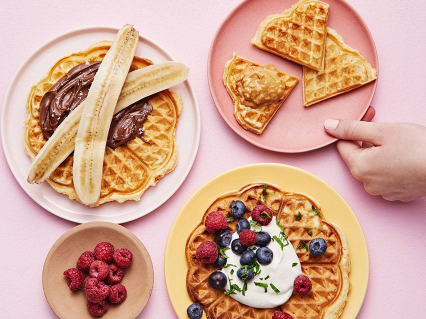 Våfflor med banan och chokladkräm, jordnötssmör, samt bär och grädde på rosa bakgrund.
