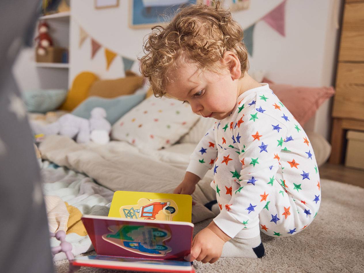 Ett litet barn i stjärnpyjamas, sittande på mattan, läser en bok.