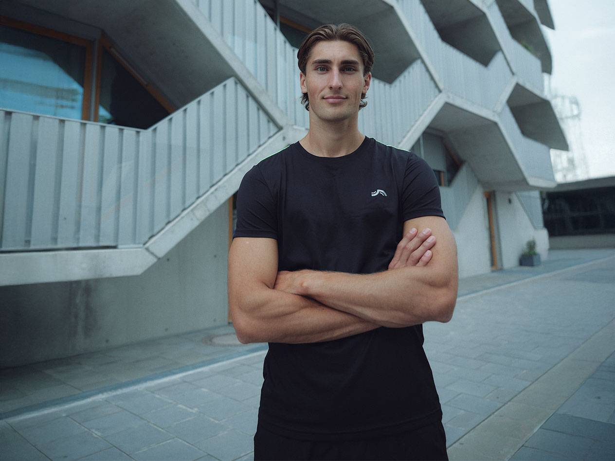 Man i svart t-shirt med logotyp och svarta shorts, står utomhus med armarna i kors.