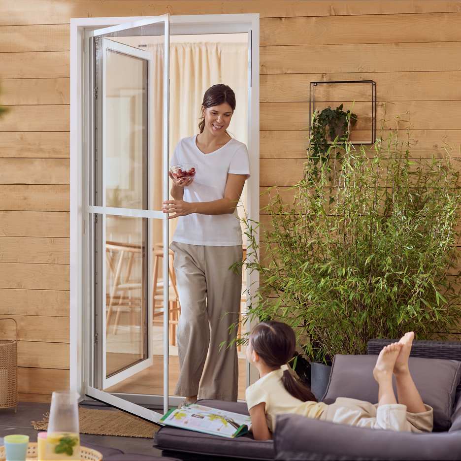 Kvinna med bär går in genom myggnät, barn läser på terrasssoffa.