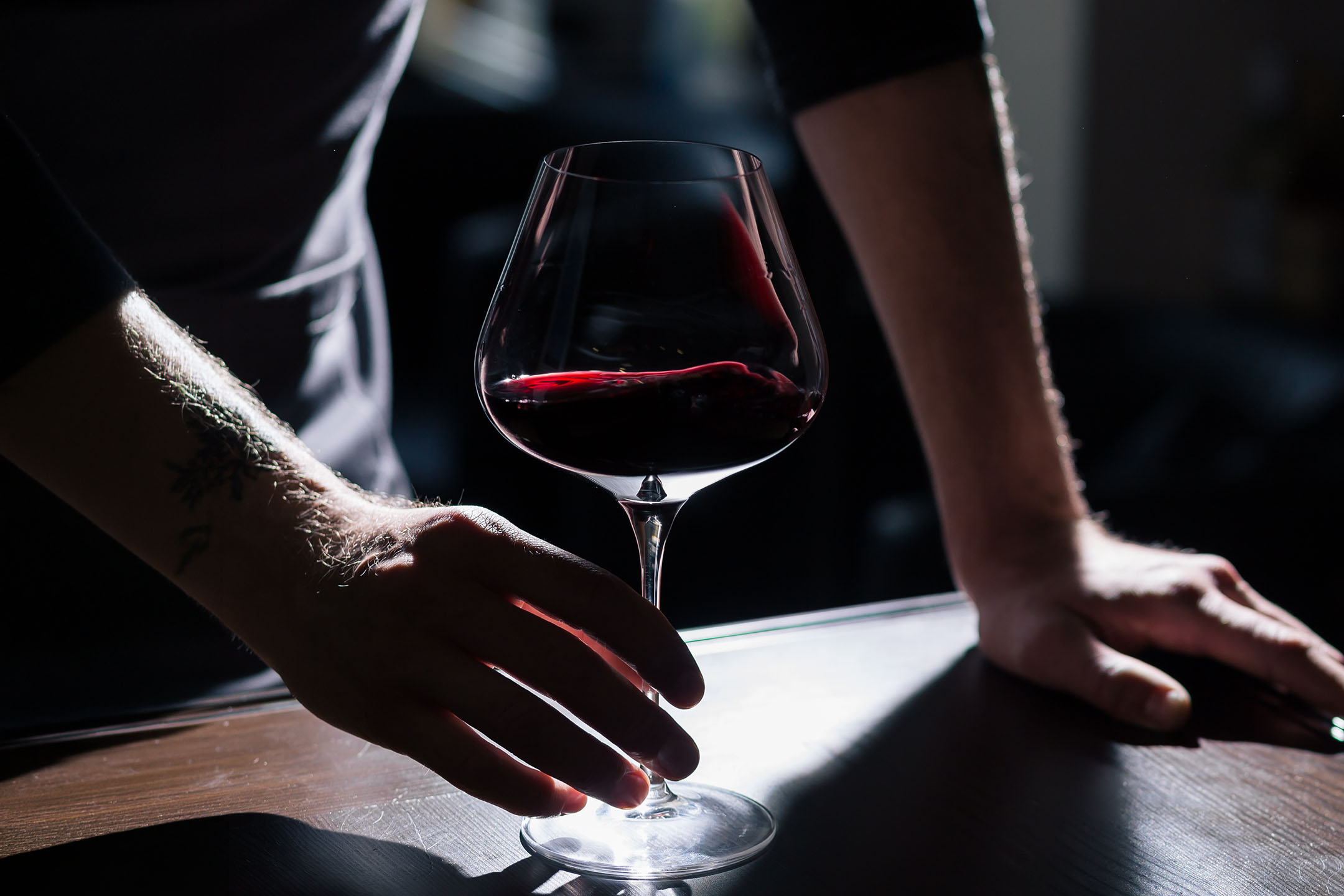 En mans tatuerade händer håller ett glas rött vin på ett träbord.