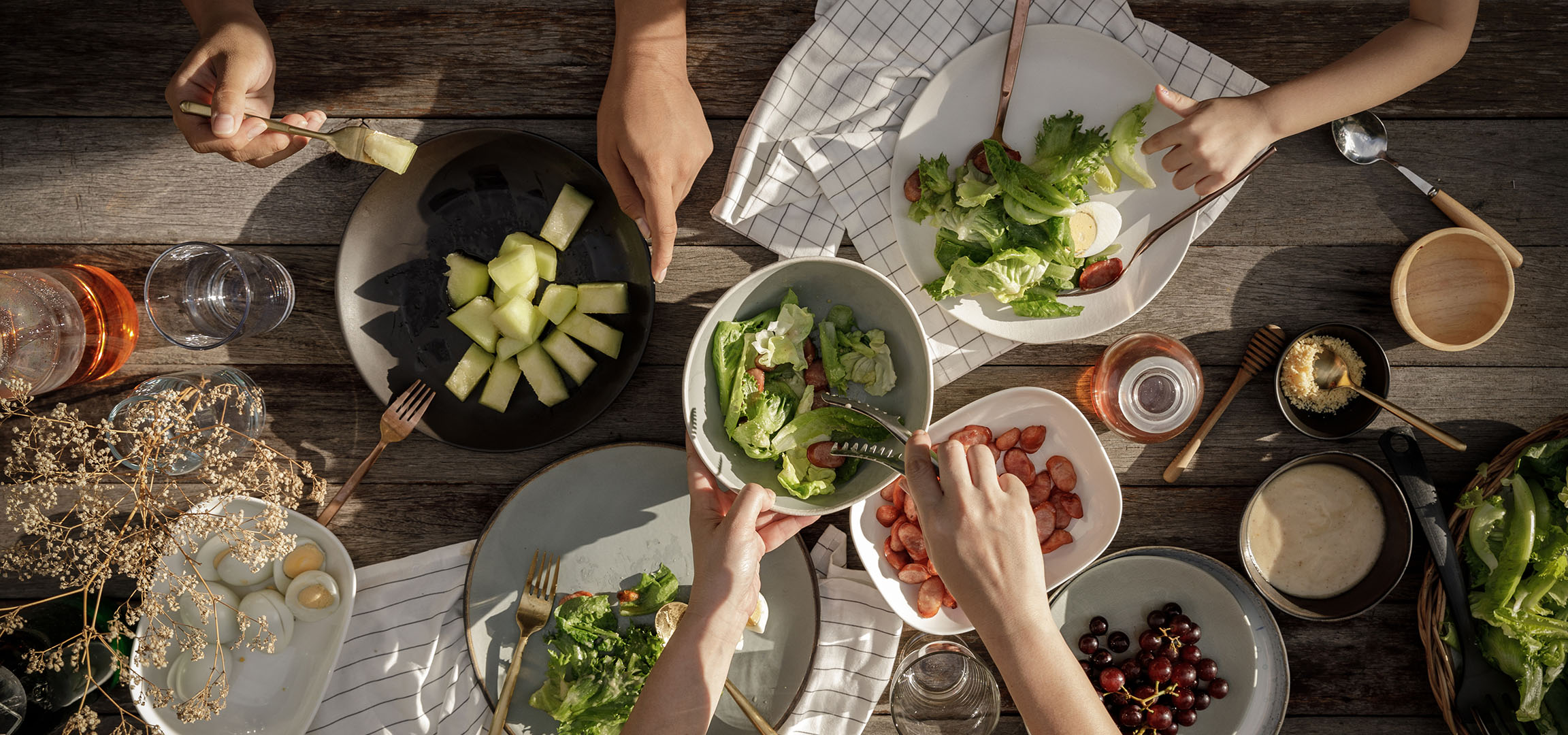 Överblick över ett bord fyllt med sallad, korv och melon, med människor som serverar mat.