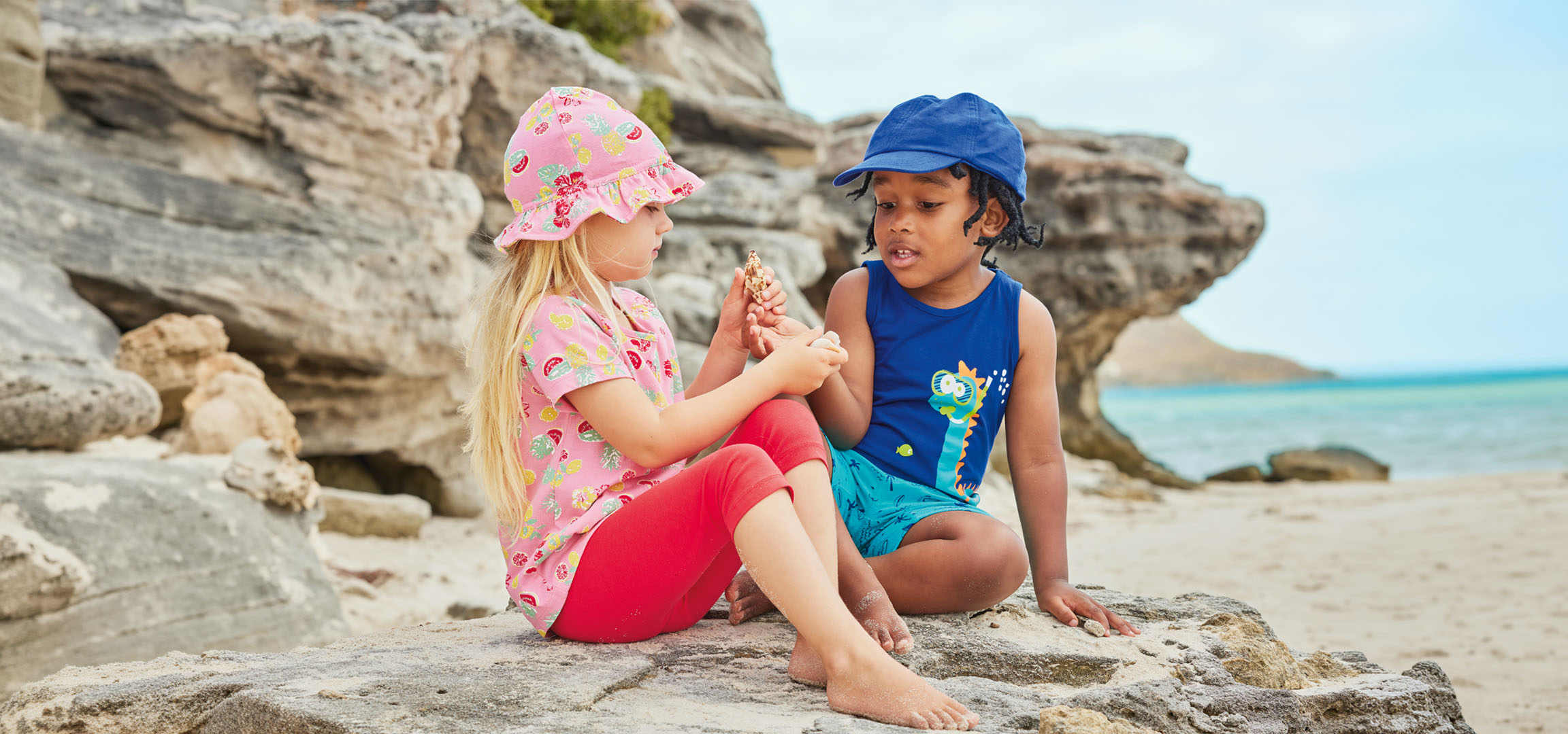 Två barn i sommarkläder sitter på en klippa vid stranden och håller i en snäcka.