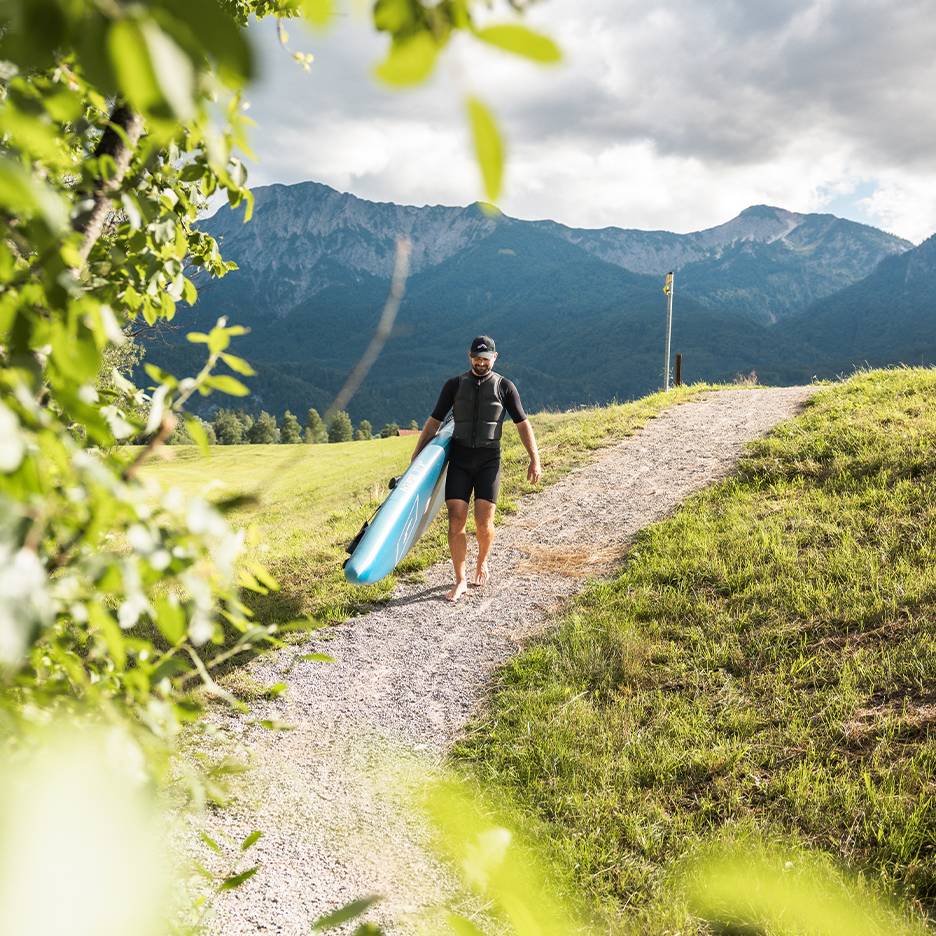 Man i våtdräkt och flytväst bär en paddleboard i ett bergslandskap.