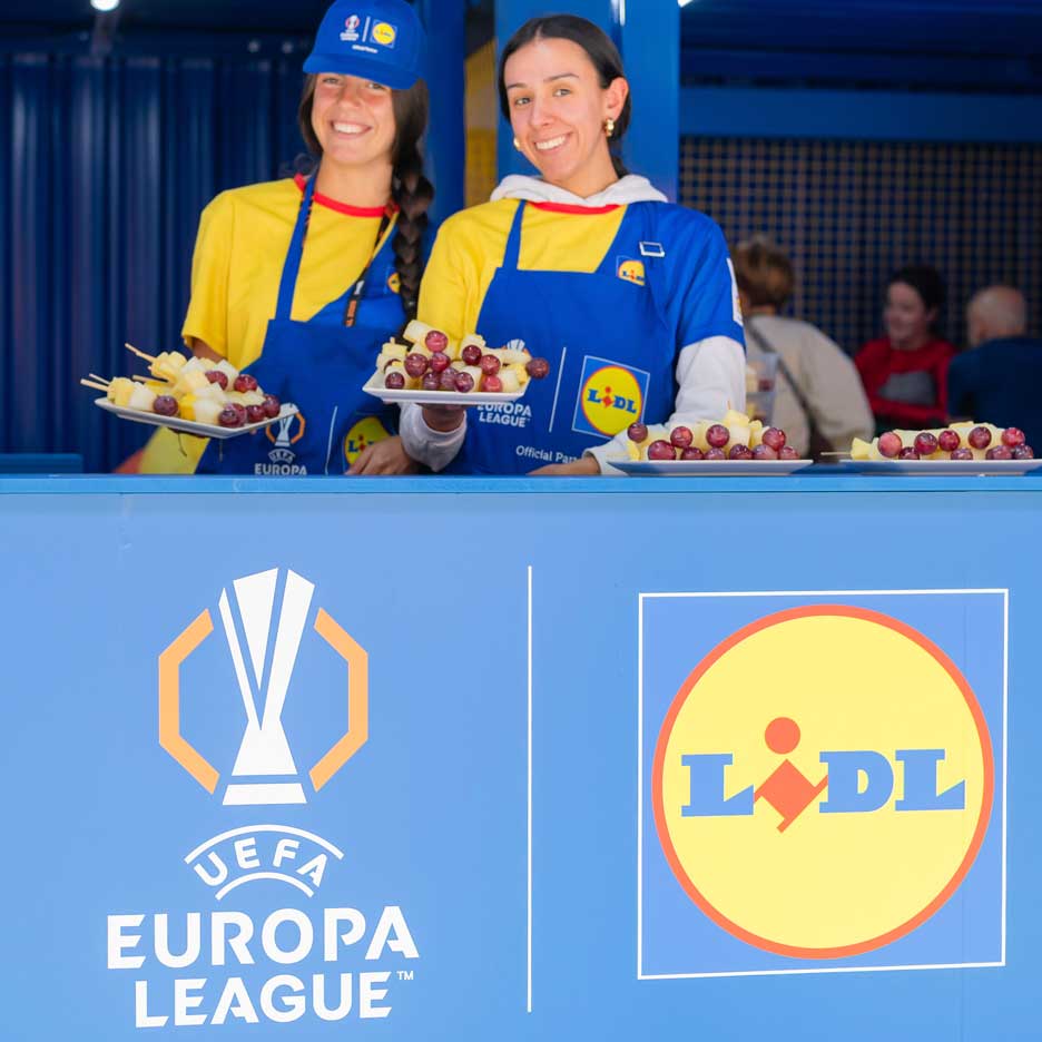 Två leende kvinnor i Lidl-förkläden erbjuder frukt på spett vid ett UEFA Europa League-evenemang.