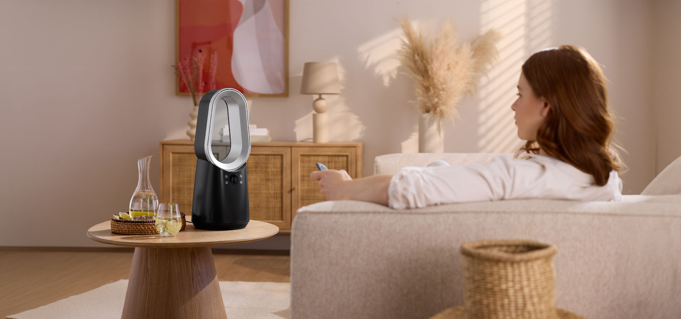 Kvinna i soffa med fjärrkontroll, modern fläkt och drycker på bordet.