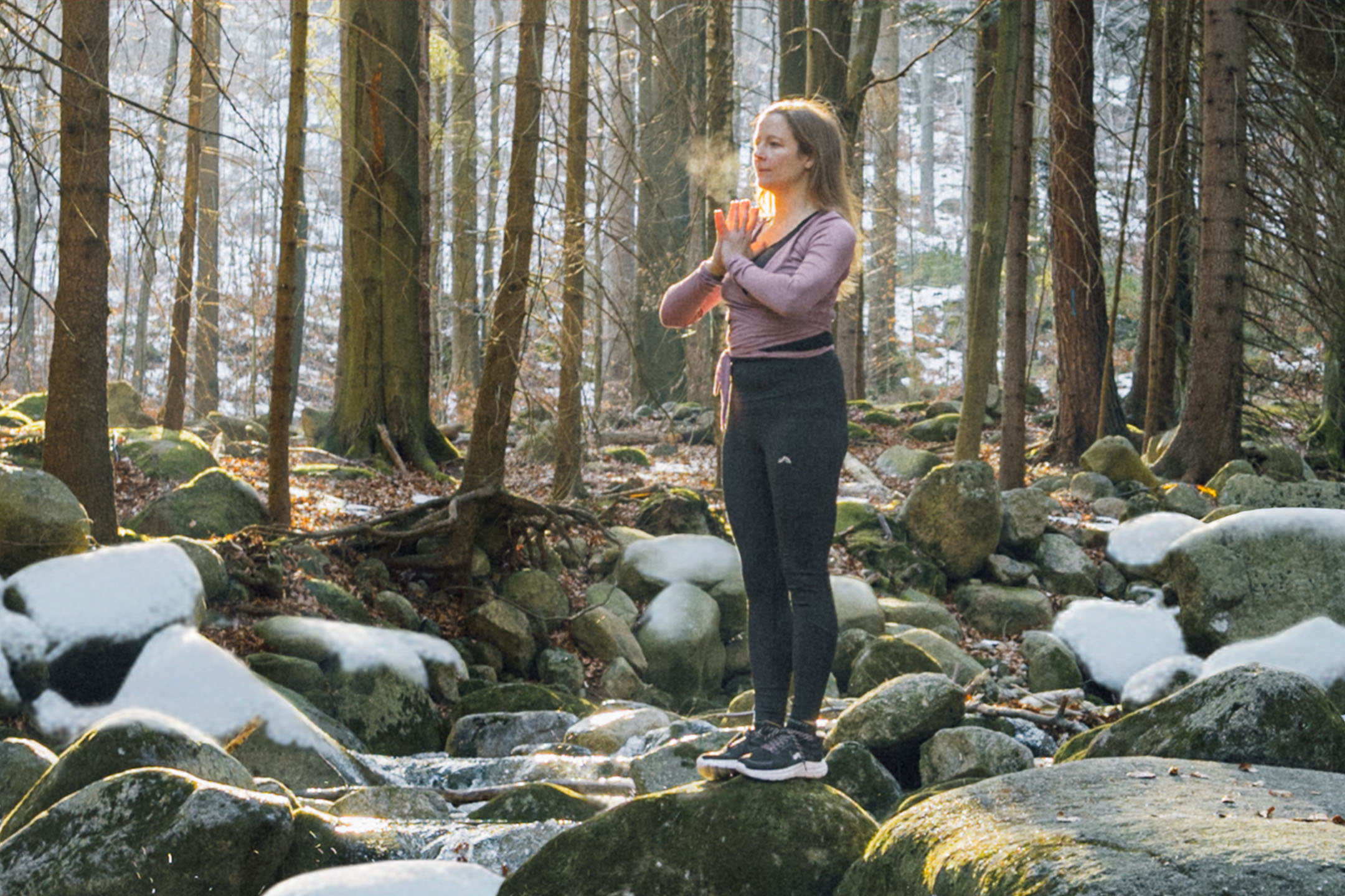 Kvinna i sportkläder som utövar yoga i en vinterskog vid en bäck.