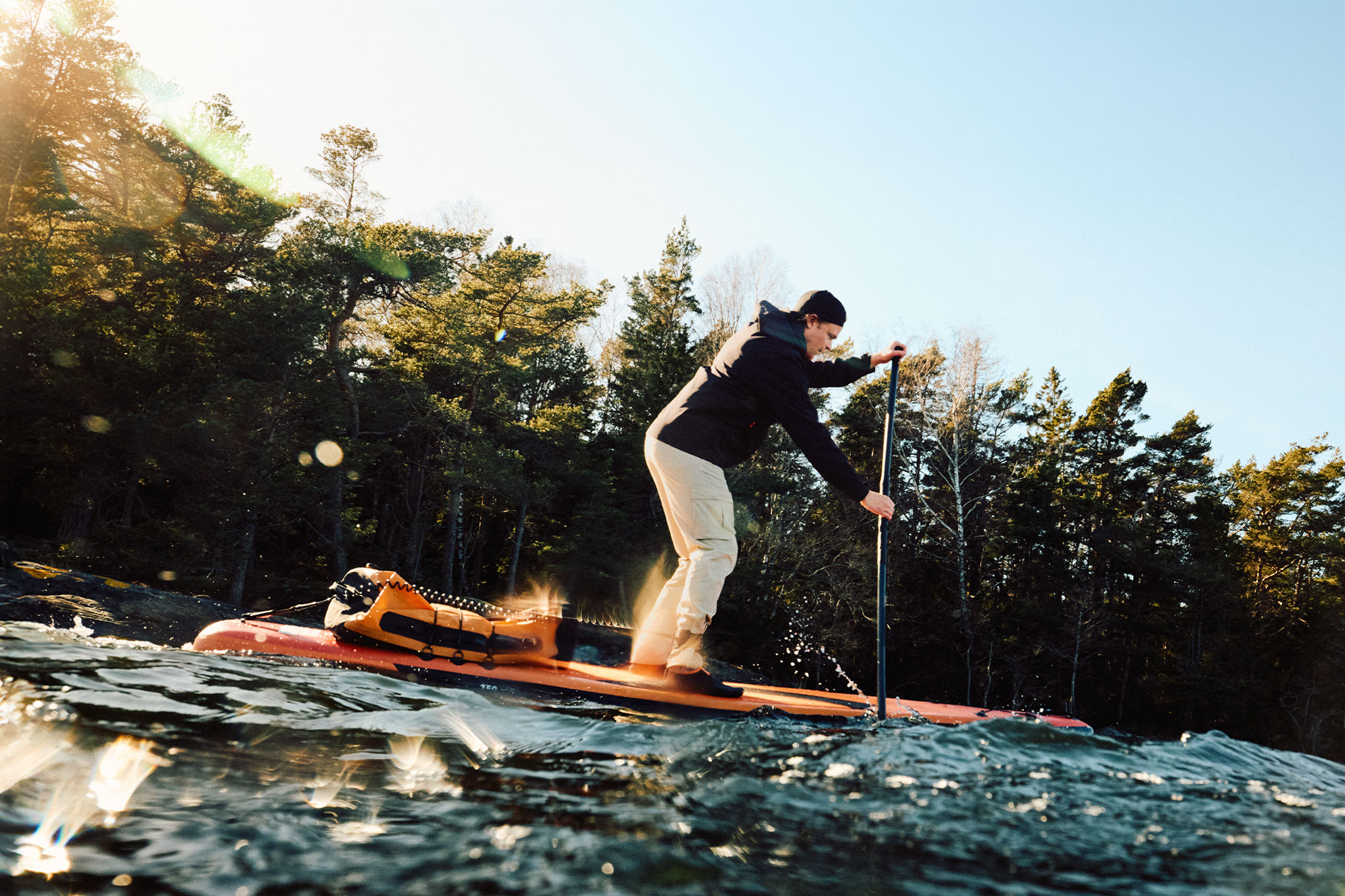 Man paddlar SUP med en orange torrväska vid en skog.