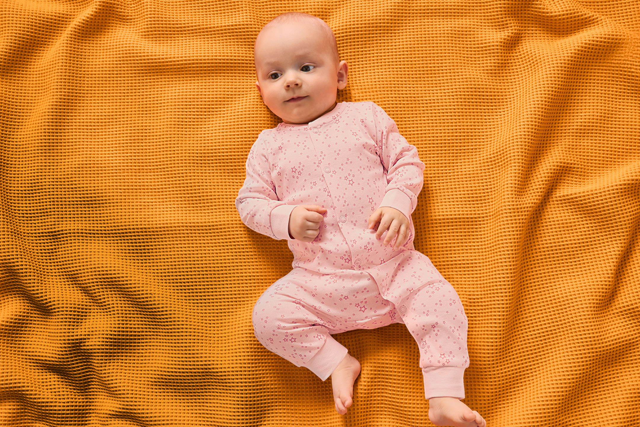 En bebis i en rosa stjärnmönstrad pyjamas ligger på en gul våfflad filt.