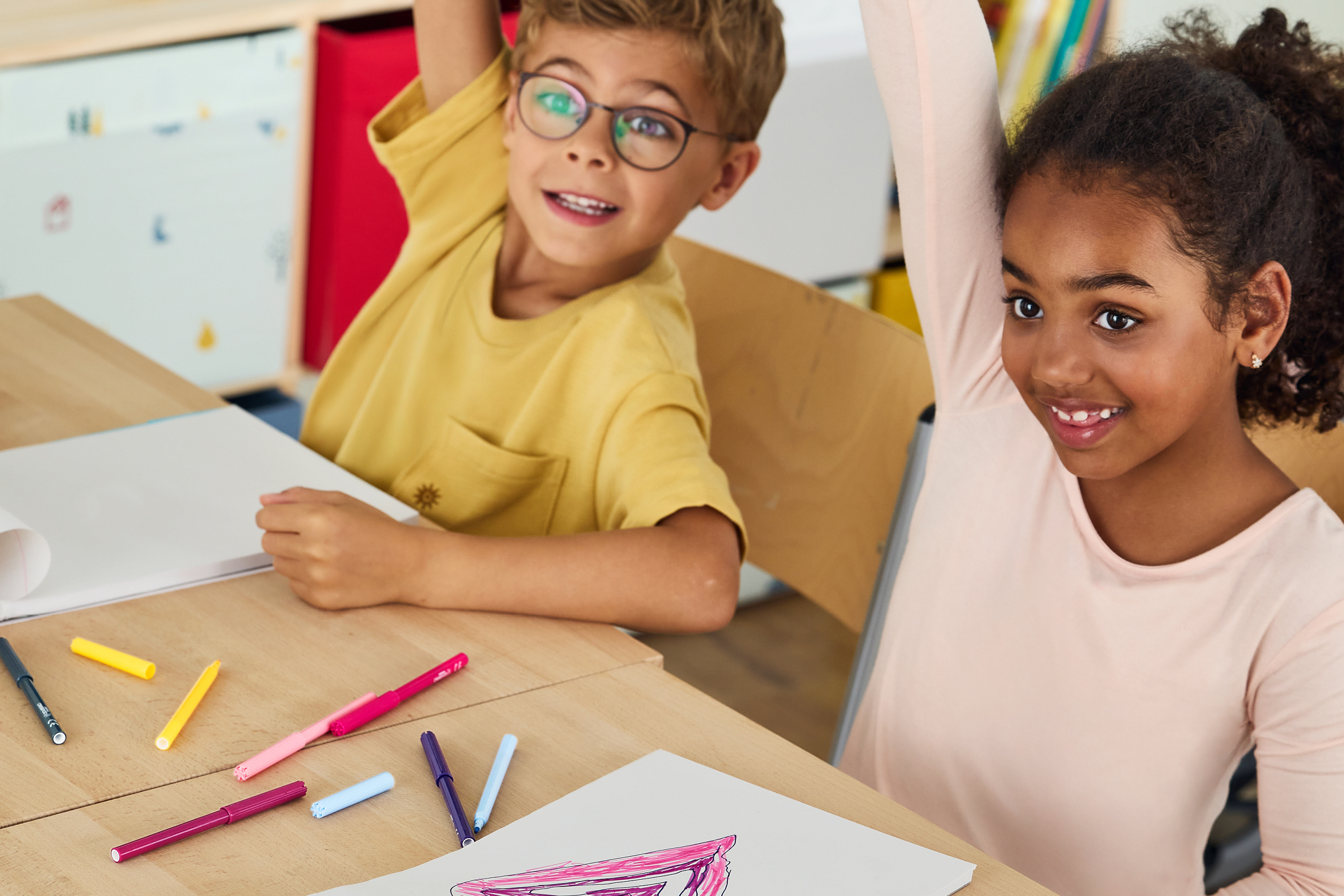 Två barn med färgpennor vid ett bord, en flicka räcker upp handen.