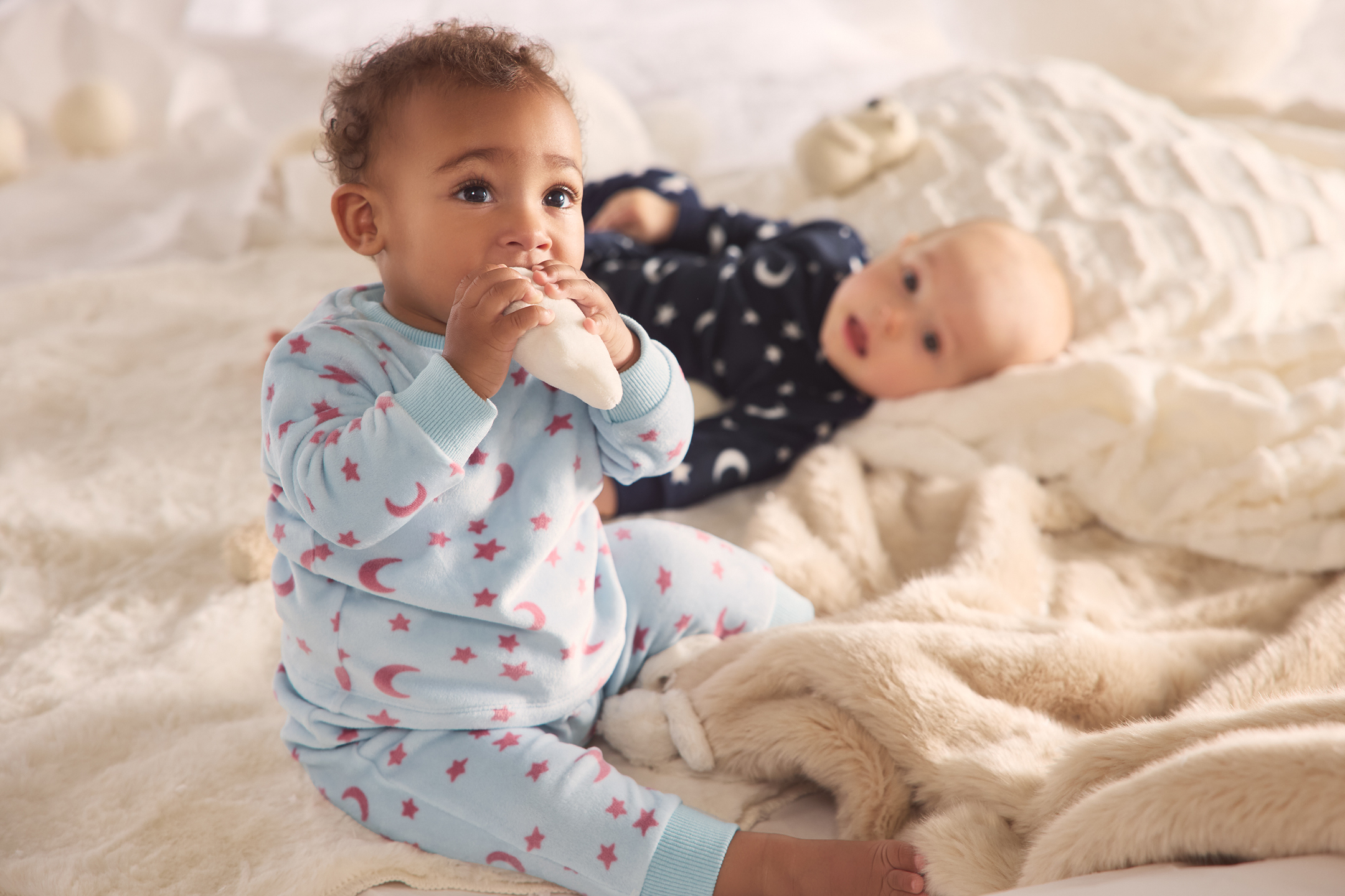 Två bebisar i pyjamas på en mysig filt, en tuggar på en leksak.