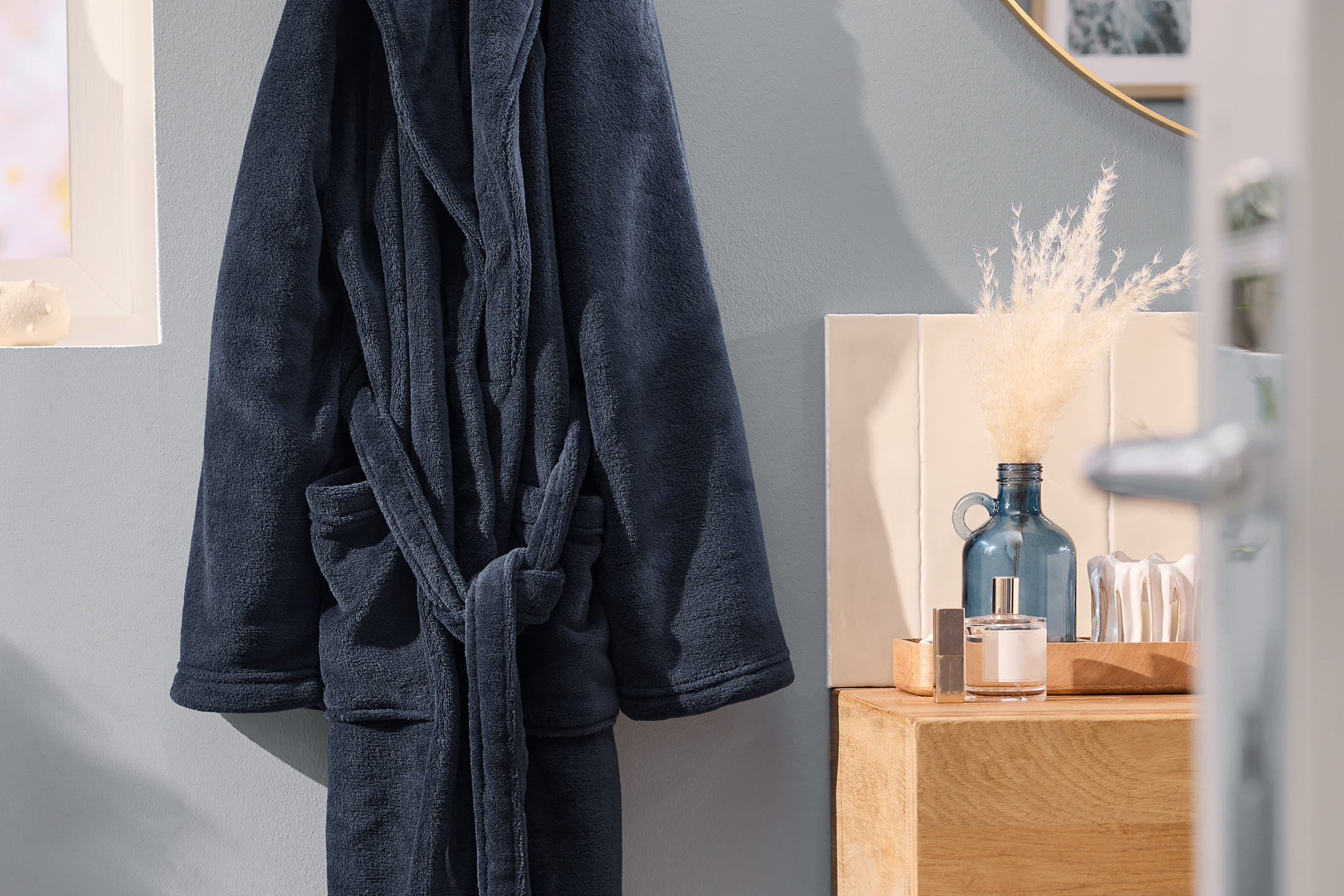 Mörkblå morgonrock hänger på en vägg bredvid en trähylla med en vas och parfym.
