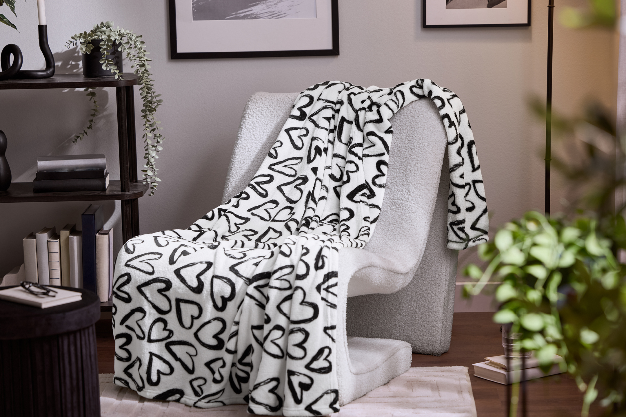 Vit filt med svarta hjärtan på en grå fåtölj bredvid en mörk hylla med böcker och växter.