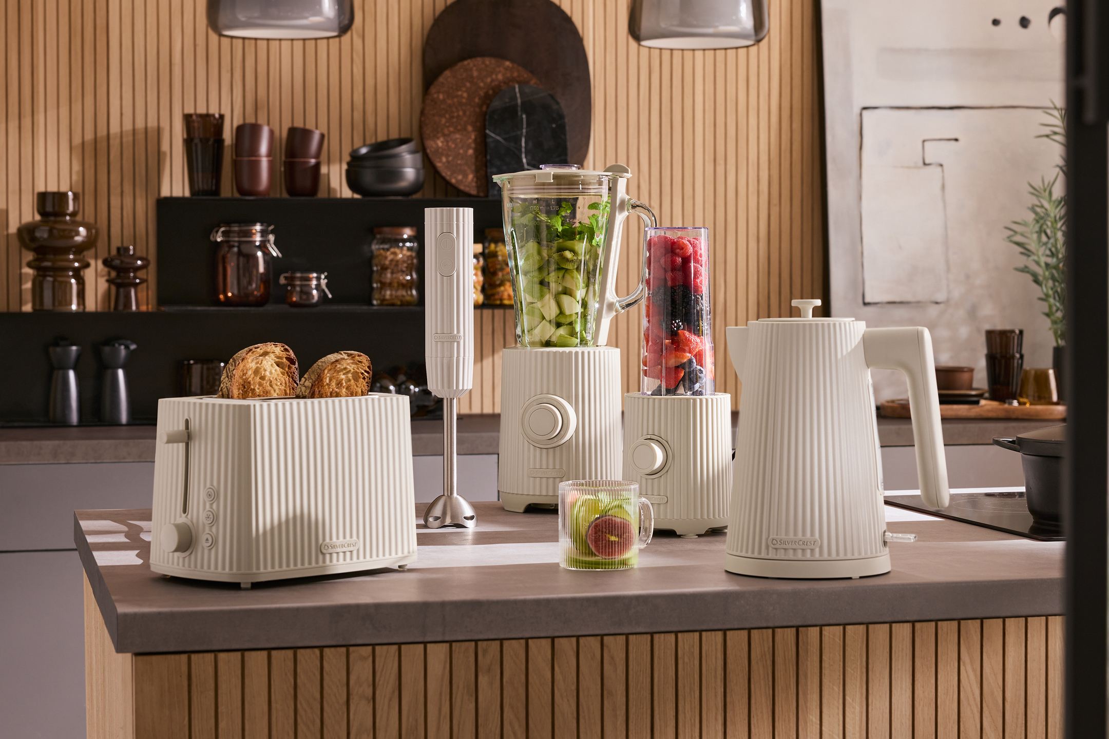 Köksbänk med beige brödrost, mixer, stavmixer och vattenkokare, med frukt och bröd.