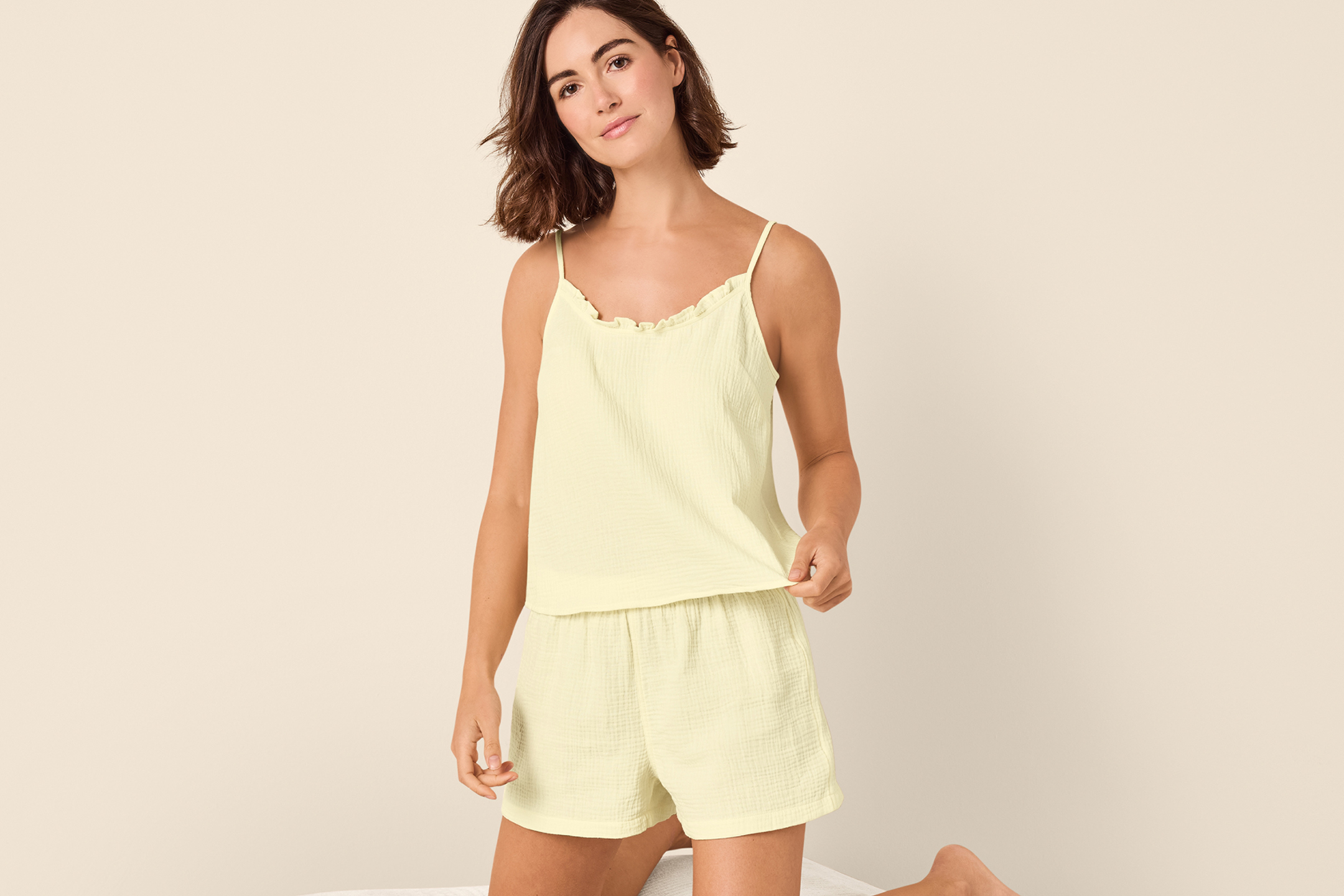 Kvinna i ljusgul pyjamas med shorts och linne, knästående på en säng.