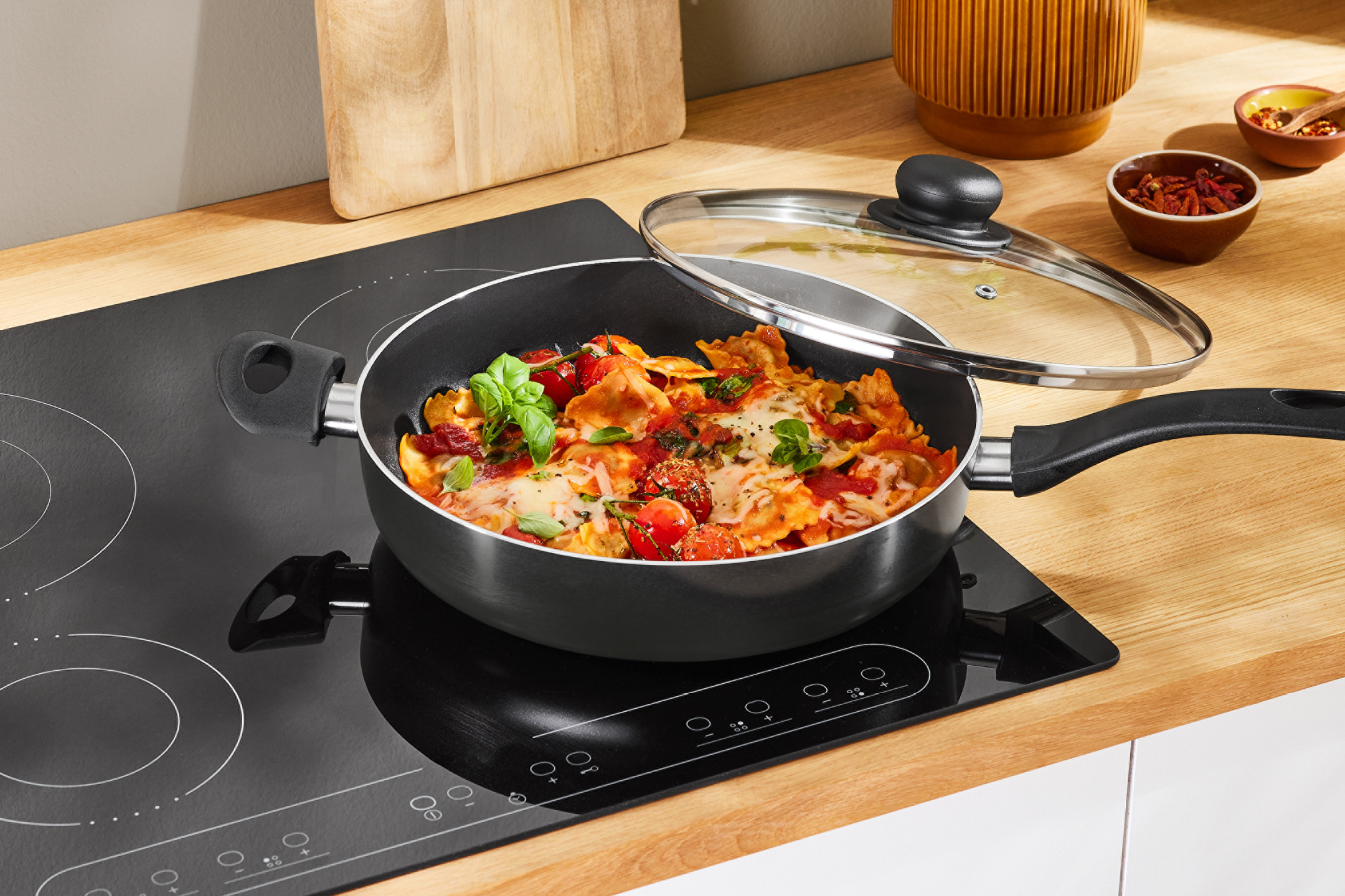 Stekpanna med ravioli på en induktionshäll, med träskärbräda och kryddskålar i bakgrunden.