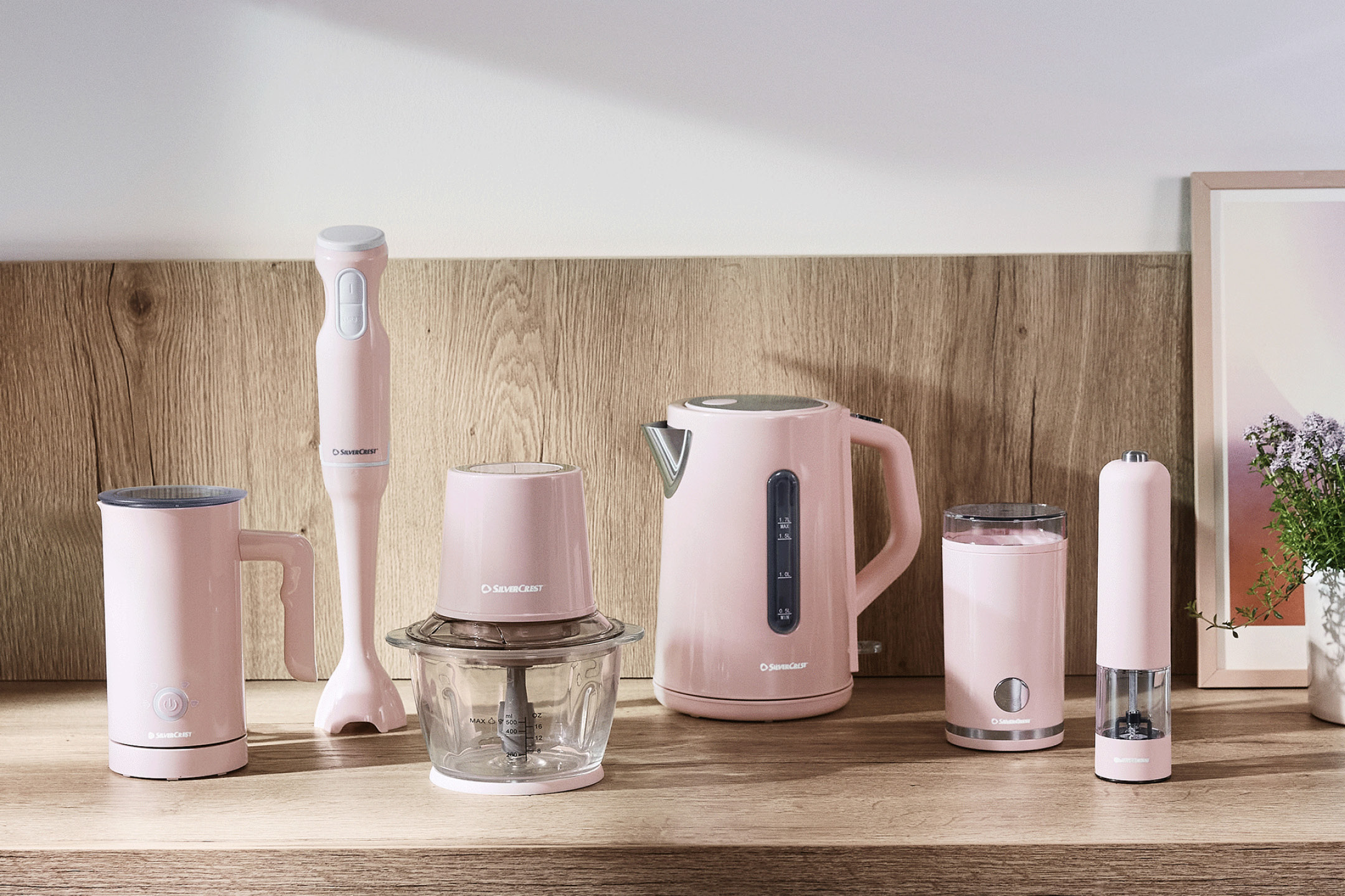 En rad rosa köksmaskiner, inklusive en stavmixer, matberedare, vattenkokare och mjölkskummare, på en träbänk.