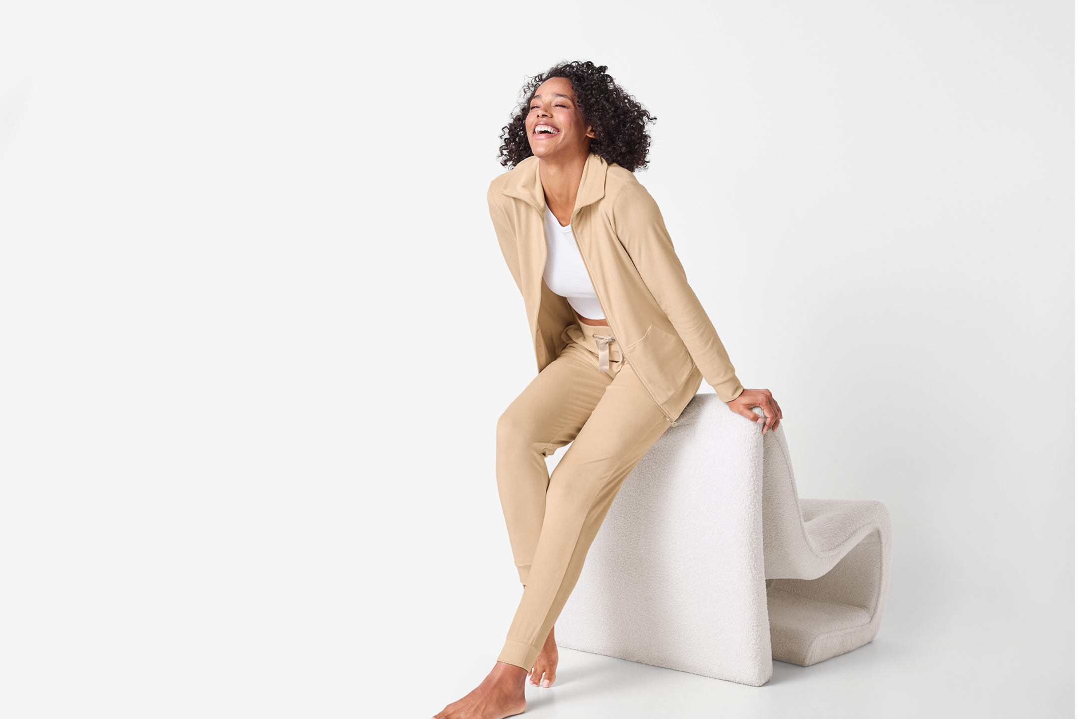 Kvinna i beige träningsset och vit topp sitter på en vit stol och skrattar.
