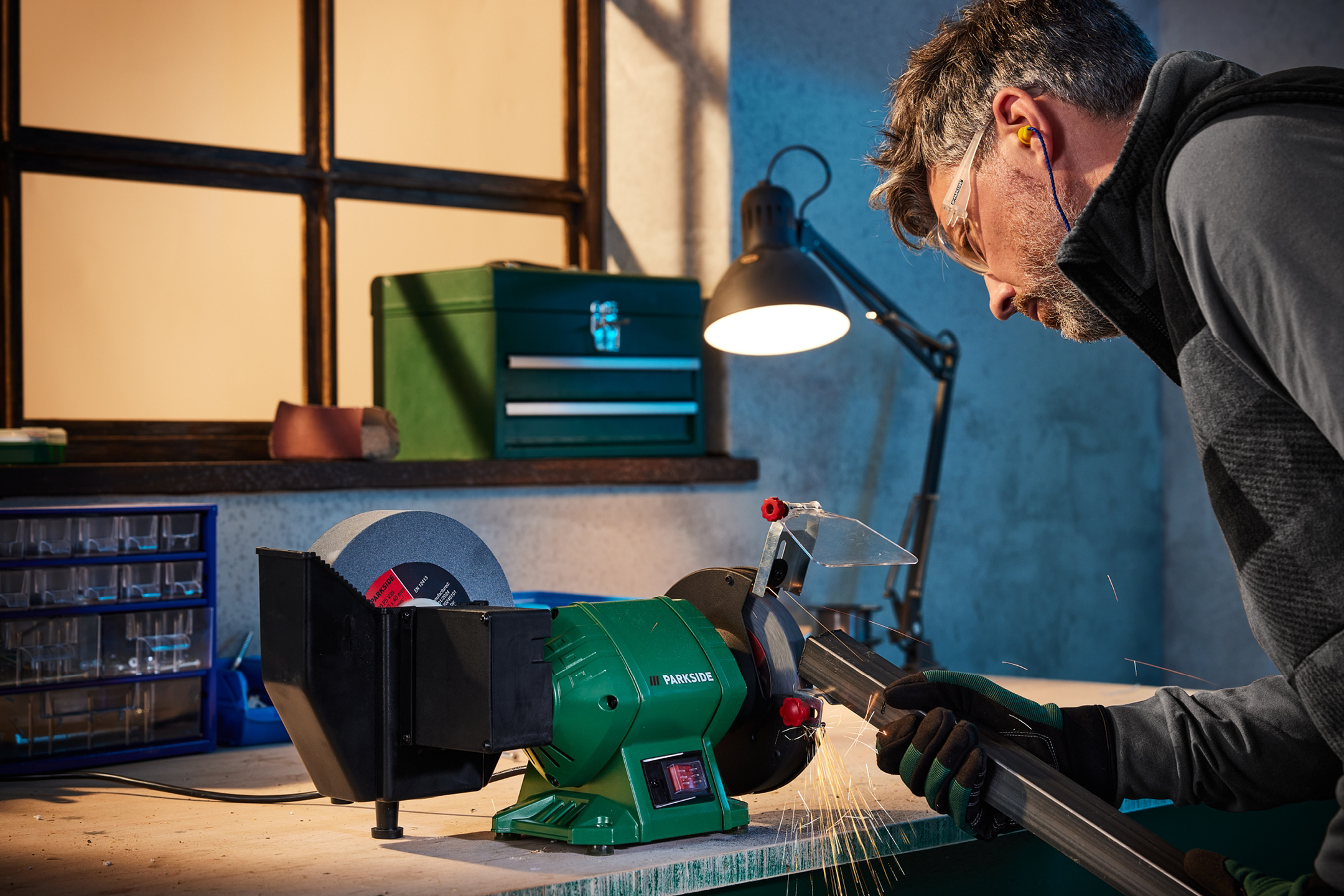 Man slipar metall med en Parkside bänkslipmaskin, med skyddsglasögon och hörselskydd.