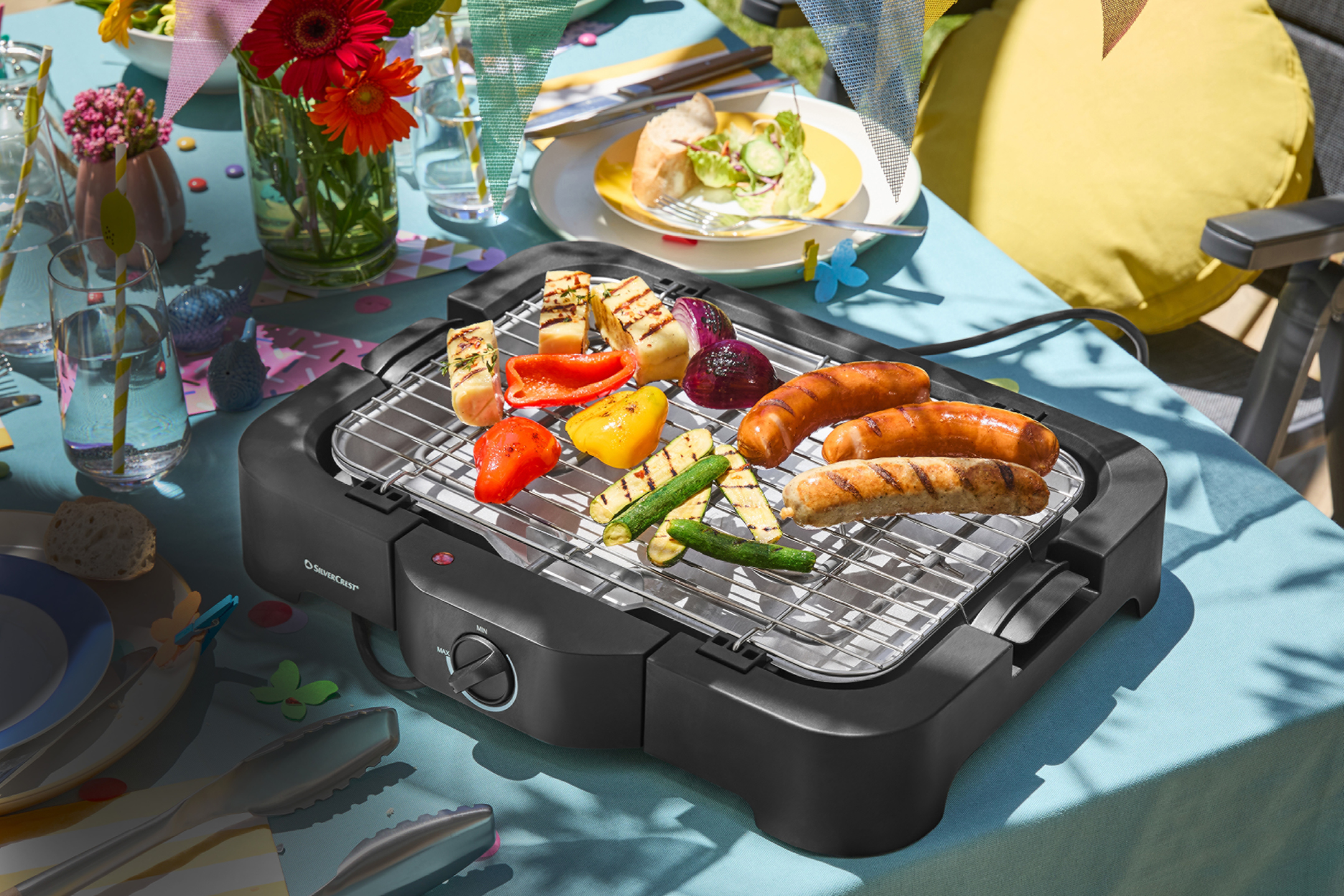 Elektrisk grill med korv och grillade grönsaker på ett dukat bord utomhus.