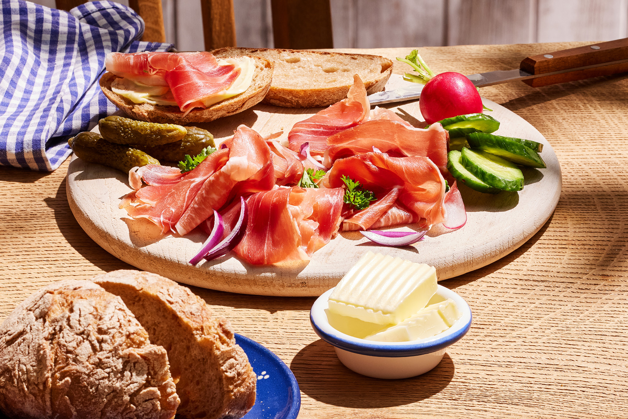En träskiva med skinka, saltgurka, smör, bröd och ett glas öl på ett träbord.