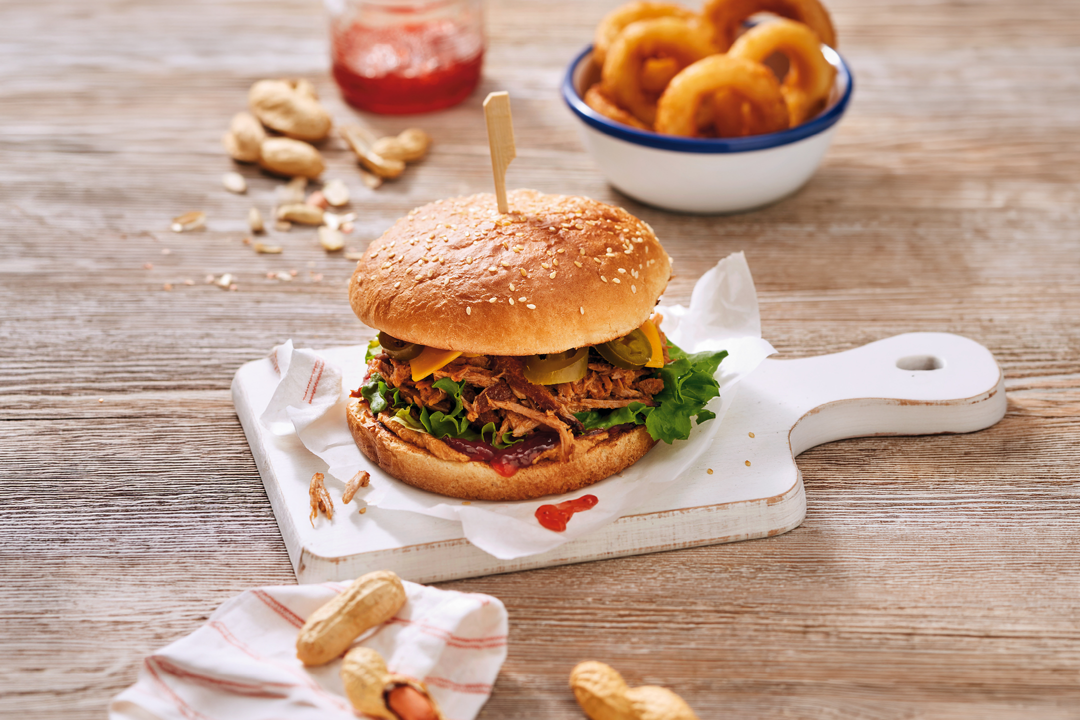 Pulled pork-burgare med ost, jalapeños, sallad, lökringar och jordnötter på träbord.