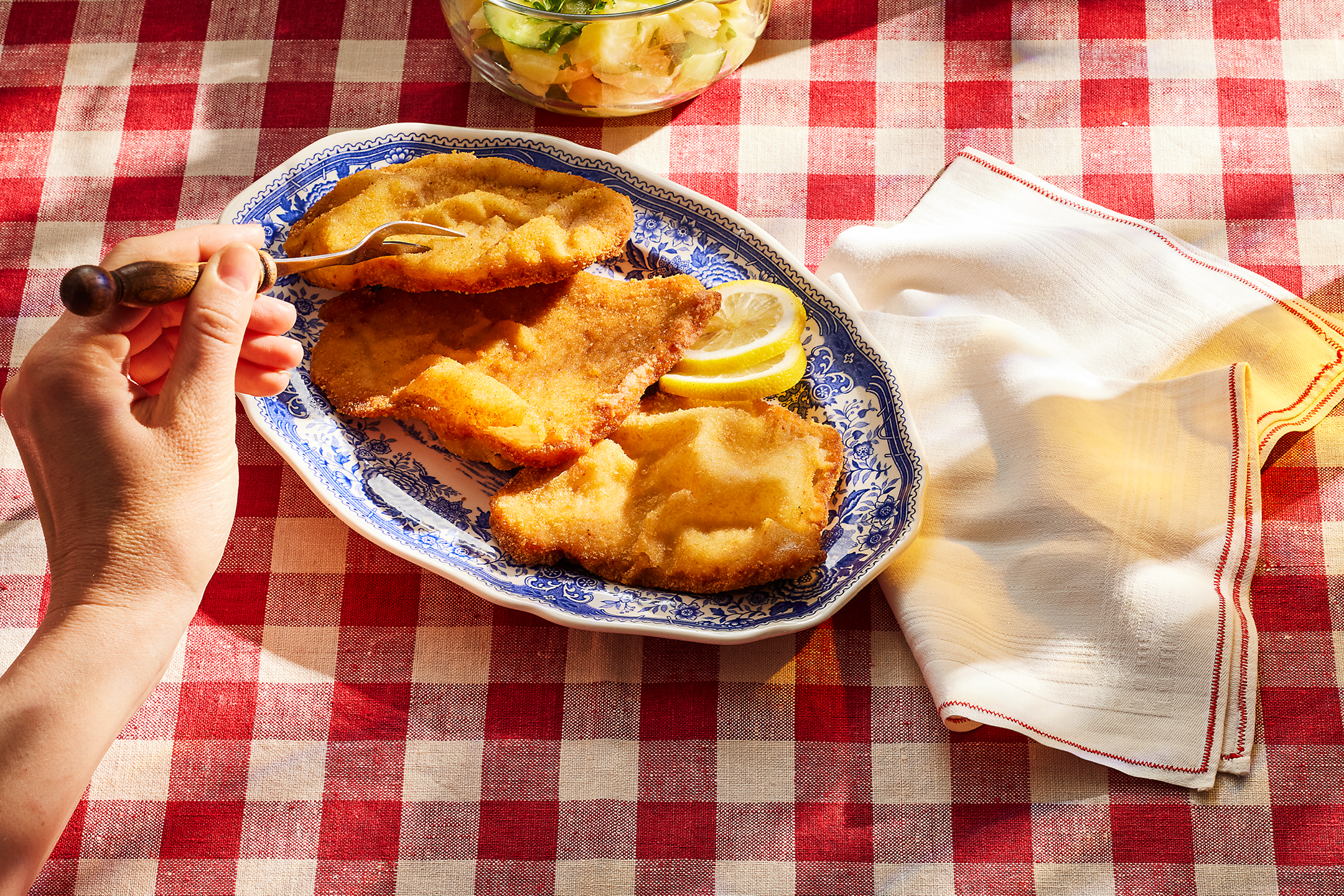En hand håller en gaffel över panerad schnitzel med citron på en rutig duk, med sallad i bakgrunden.