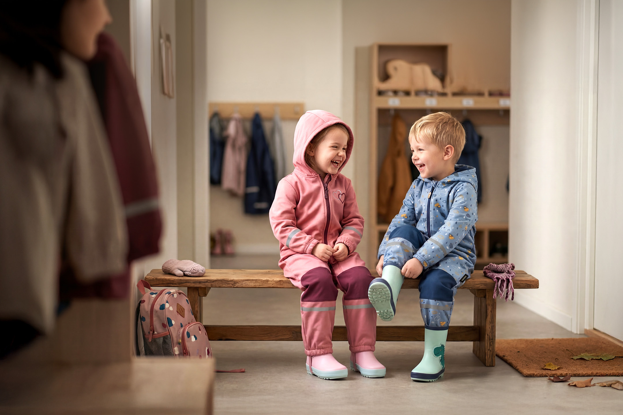 Två glada barn i rosa och blåa overaller sitter på en träbänk i en hall.