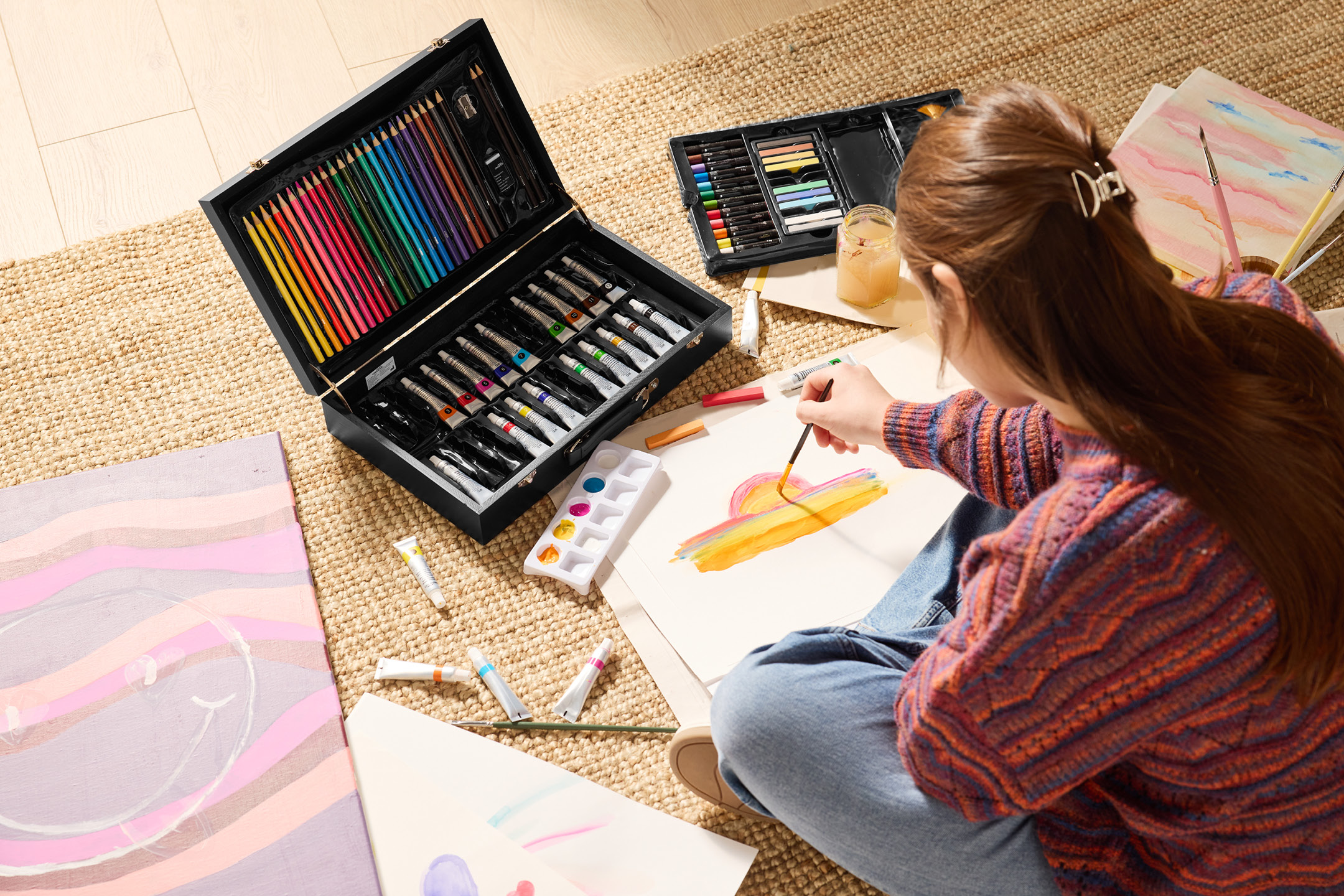 En person målar med pensel och akvarellfärger, omgiven av ett konstnärsset med pennor och tuber.