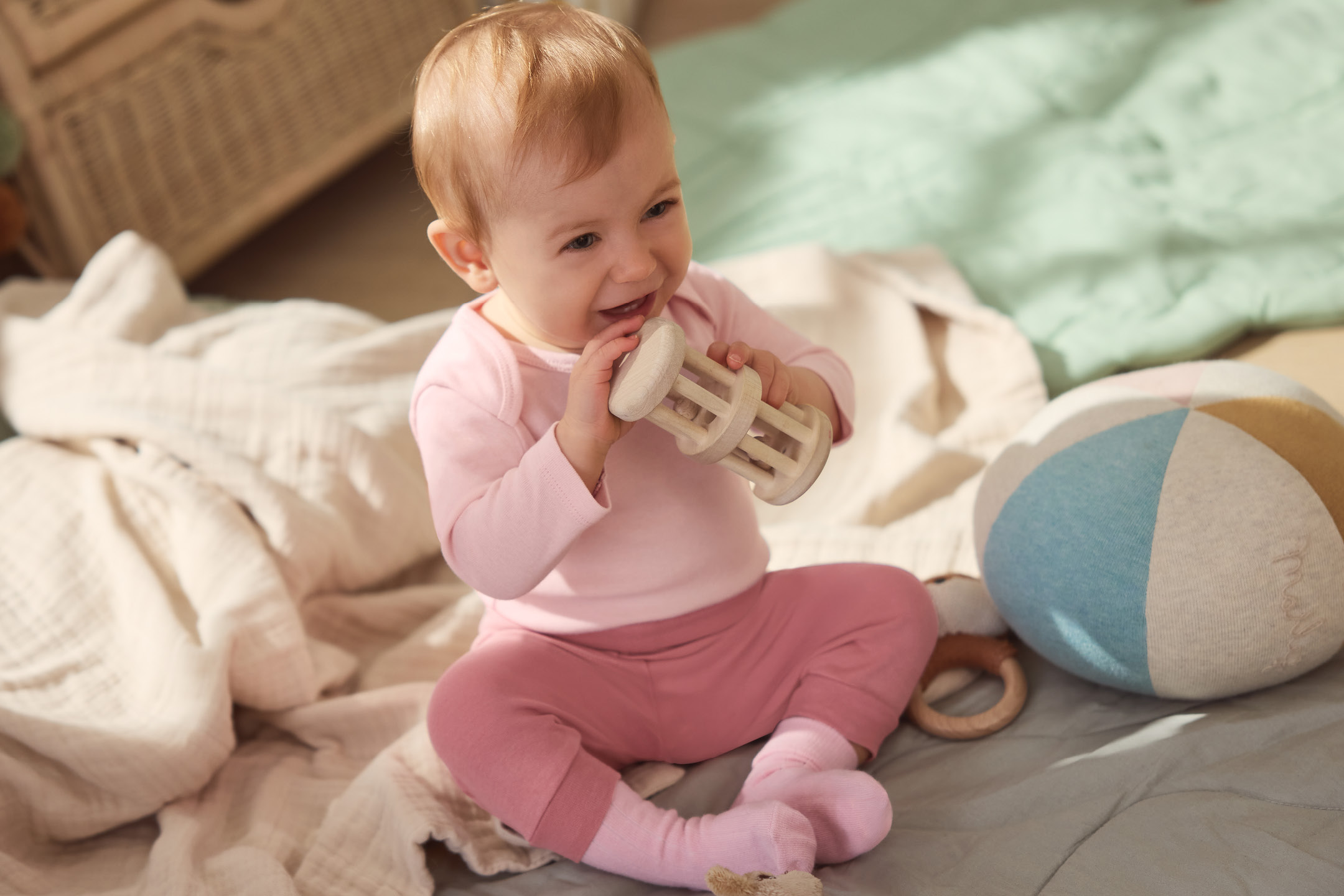Glad bebis i rosa kläder leker med en träleksak på en filt.