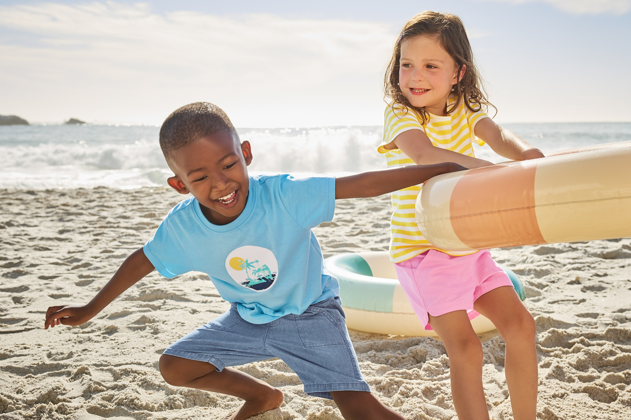 Två barn leker på stranden med badringar, klädda i somriga t-shirts och shorts.