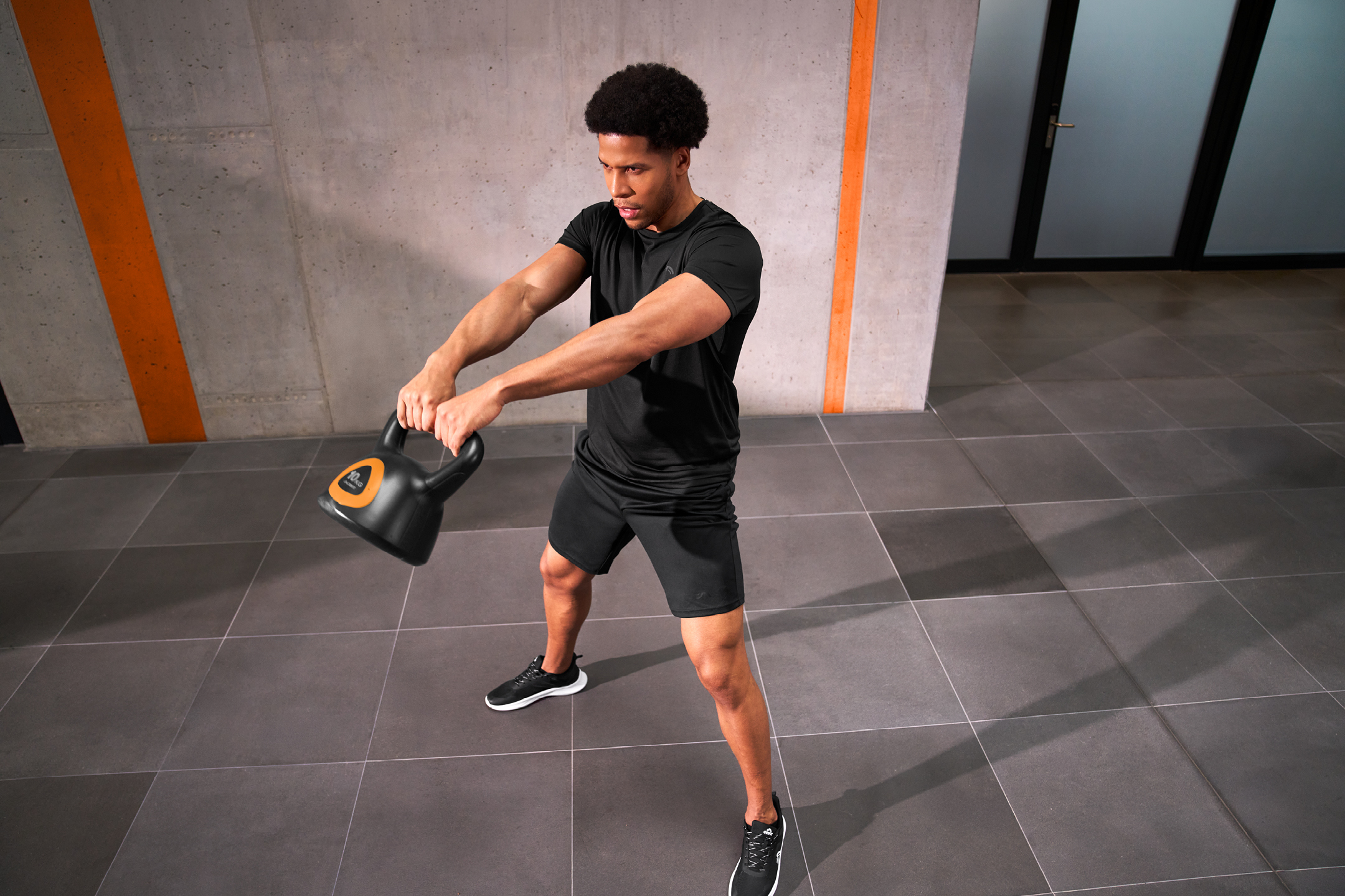 Man tränar med en svart kettlebell i ett gym med betongväggar och orangea ränder.