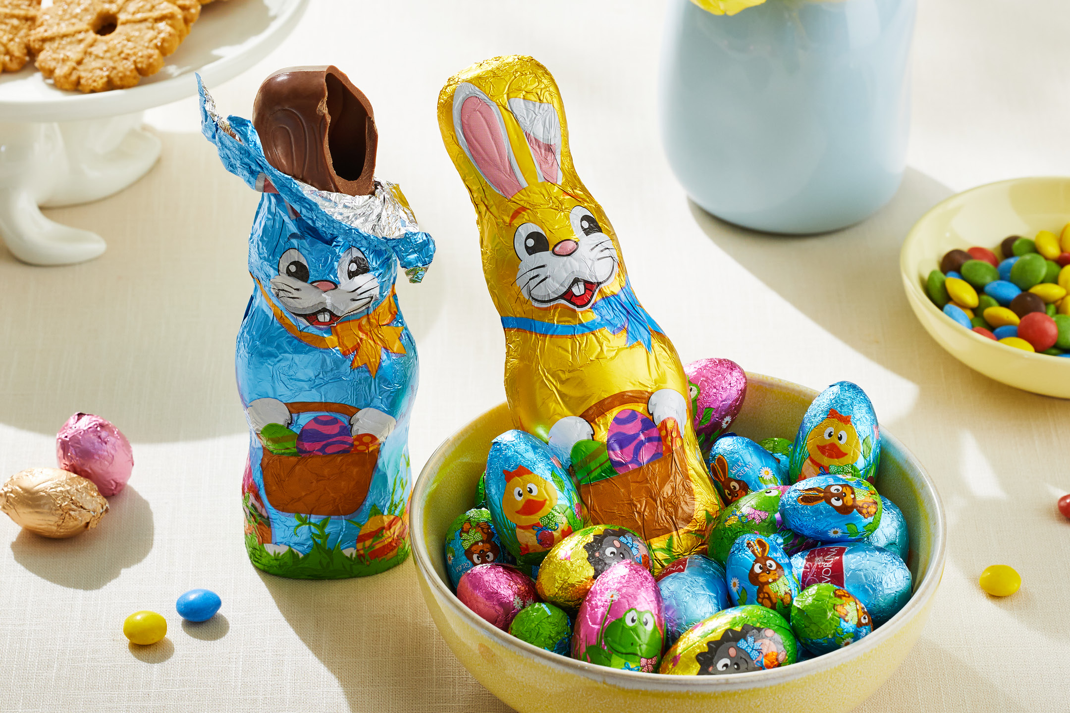 Påskgodis med chokladharar och folieinslagna ägg i en skål, samt kakor i bakgrunden.