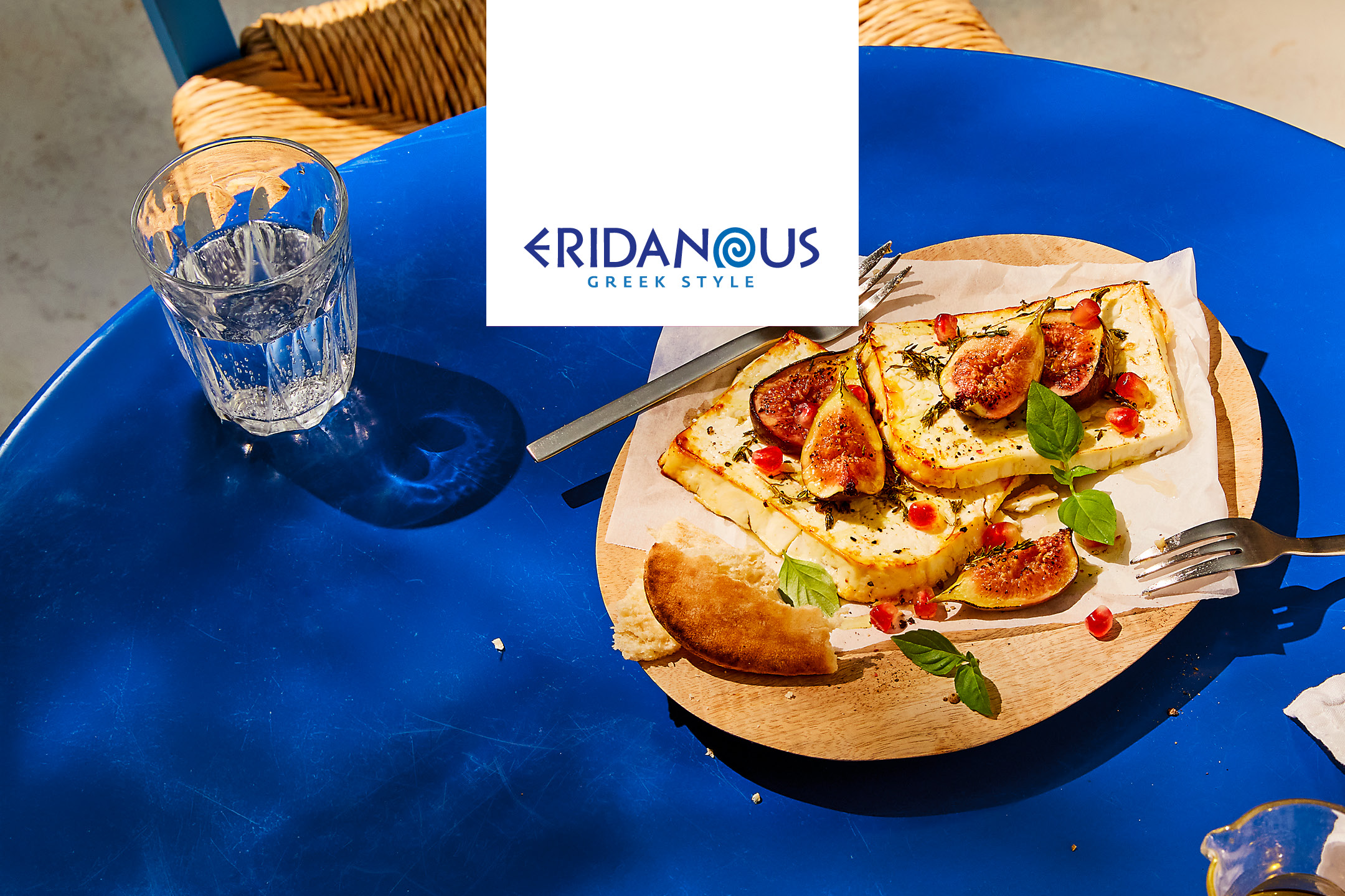 Grillad ost med fikon och granatäpple på ett träfat, ett glas vatten på ett blått bord.